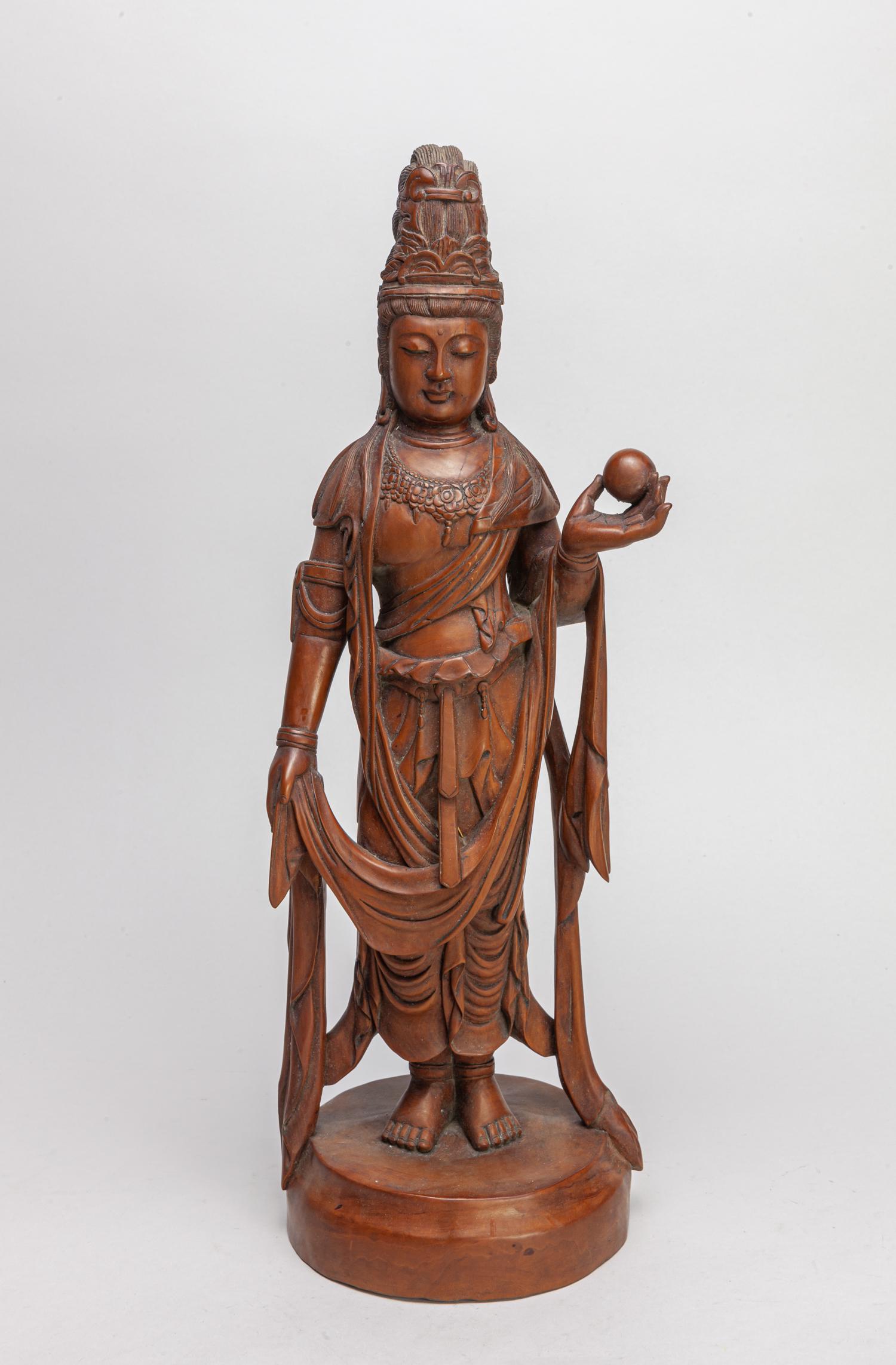 Chinese Boxwood Carved Figure of Kuanyin: The standing figure wearing a long flowing robe, with her left hand holding a sphere and right hand carrying a shawl, her hair tied up, with a round base. 17.2inch H, 5.5inch W.