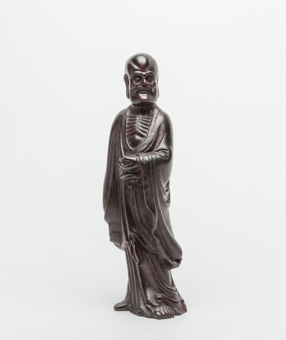 Chinese Zitan Like Wood Carved Luohan: Carved standing Luohan wearing a loosely fitting robe opening at the chest, the left hand holding a fruit, the face with a serene expression, moulded with long eyebrows, a pronounced cranium, and a pa