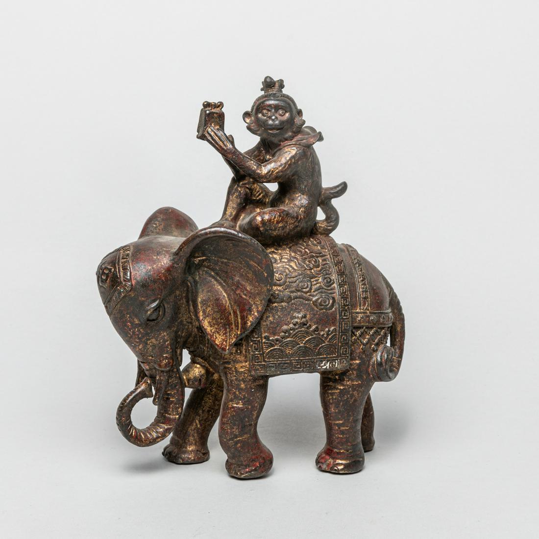 Collectible Chinese Gilt Metal Table Sculpture: Cast as a standing elephant decorated with a Xiang character at its forehead and a rug carved with clouds and waves, a monkey seated on its back with head tuned to the right and hands holding a wrappe