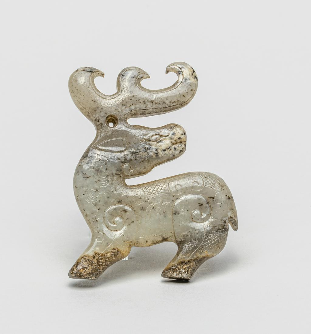 Chinese Jade Carving of Deer: The flattened carving modeled in deer form, the head turned sharply backwards, detailed with curling antler and pierced with an aperture, the mouthed gray stone with dark inclusions and calcified patc