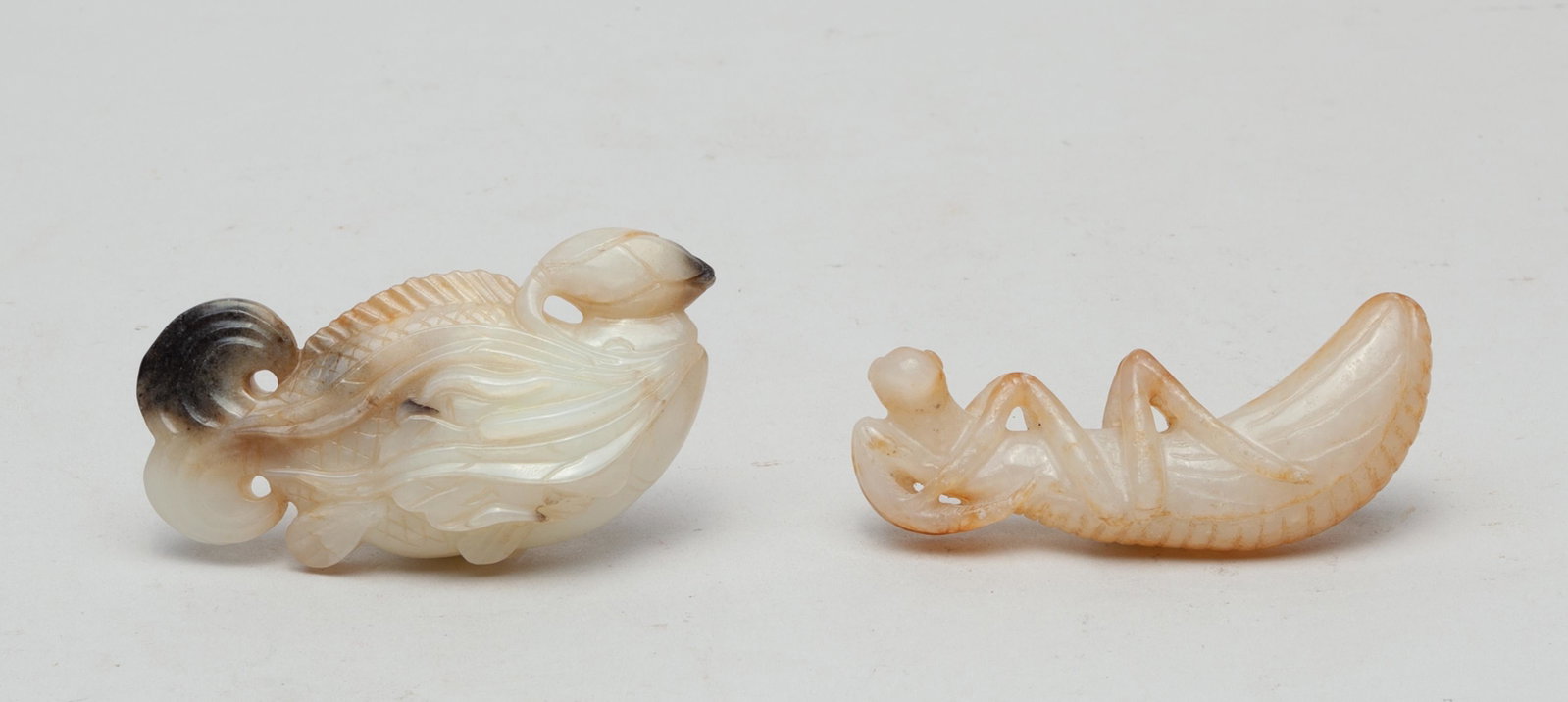 Collectible Chinese Jade Carvings: One carved as a grasshopper, the other as a fish with a bifurcated fishtail, its mouth grasping water plants and a lotus stem issuing a bud and a leaf, the stone in white tone with black and russet in