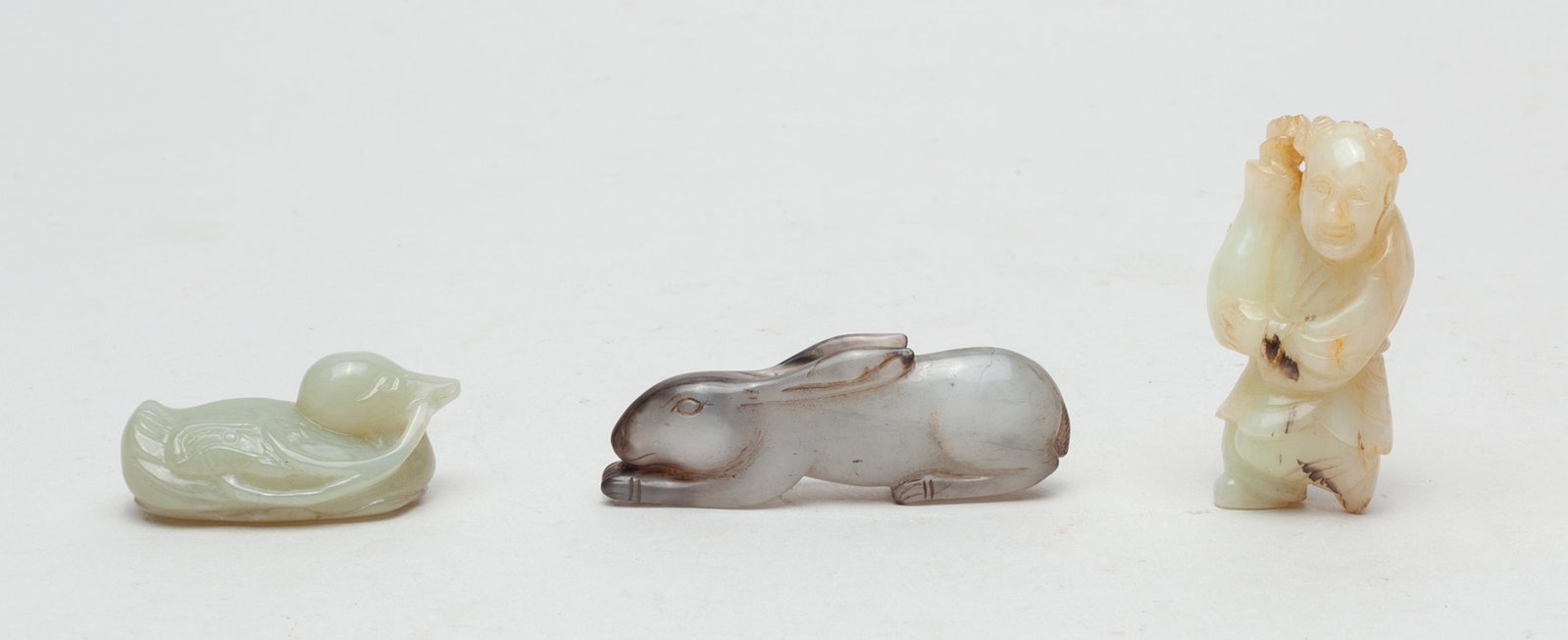 Collectible Chinese Jade Toggles: Three as a group, including a pale celadon jade duck above a lotus leaf, grasping a lotus flower stem, a black and white jade recumbent rabbit, and a russet jade figure holding a vase with flowers ins