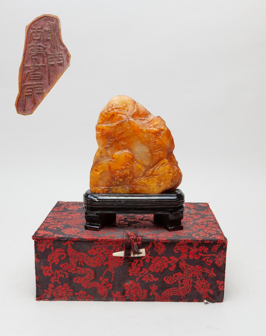 Large Chinese Stone Carved Sculpture: The honey-brown stone carved with two scholars in landscape with gnarled pine trees, craggy rockwork and a bridge, inscribed with four characters, with a wood stand and a box. 4.6inch H, 4.2inch L, 1.
