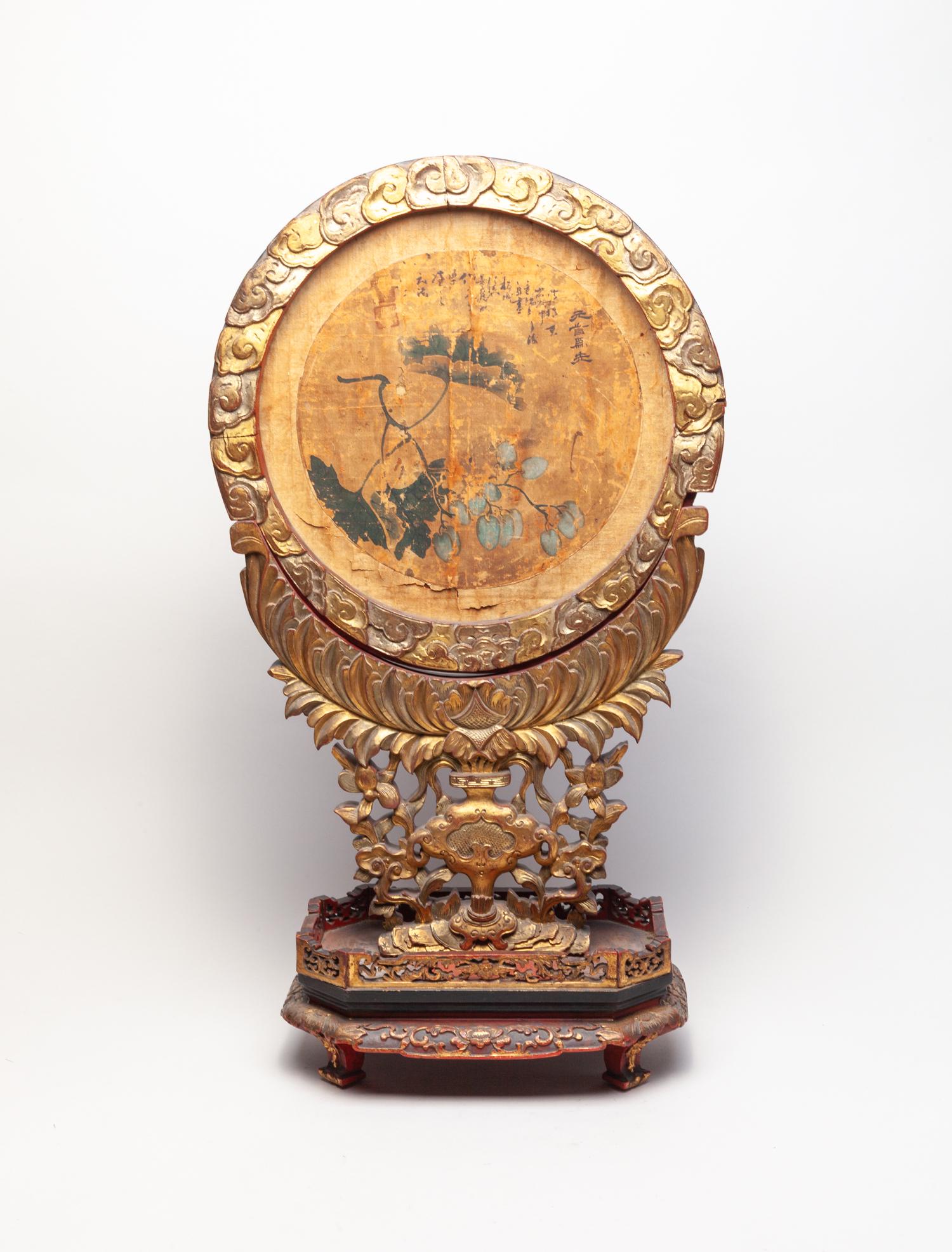 Antique Chinese Gilt Wood Table Screen: The circular gilt wood panel carved with scrolling clouds at the edge, enclosing a paper painting of fruits borne on leafy stems with inscription, the stand carved in openwork with flowers and ruyi he
