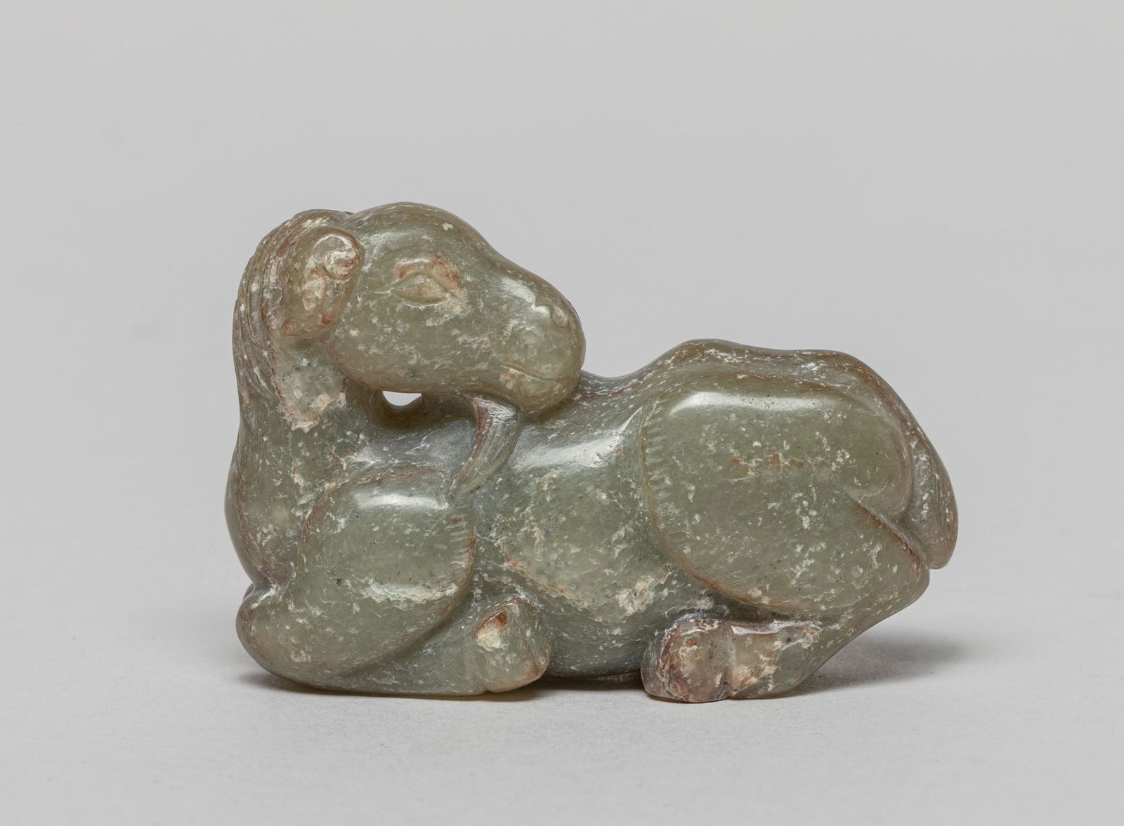 Chinese Chicken Bone Color Jade Horse: Carved in the form of a horse recumbent with legs tucked beneath its body and head turned sharply backwards, the stone in a mottled celadon tone with opaque inclusions. 2.3inch L, 0.75inch W, 1.5inch