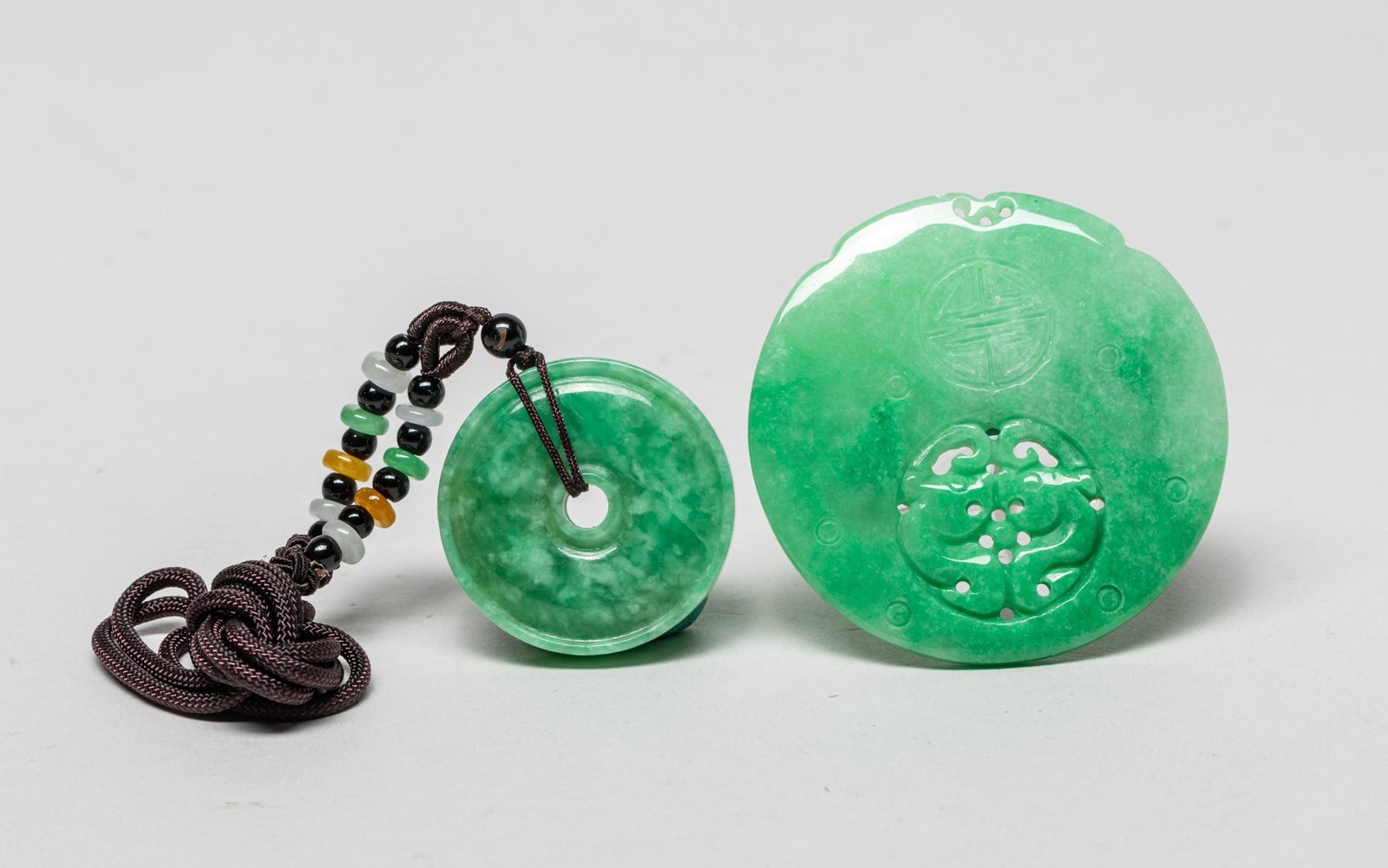 Two Chinese Jade Jadeite Like Pendants: Both of circular shape, one pierced with an aperture at the centre, the other carved in openwork with a pair of confronting bats, beneath stylized shou characters, the stone in a mottled apple green t