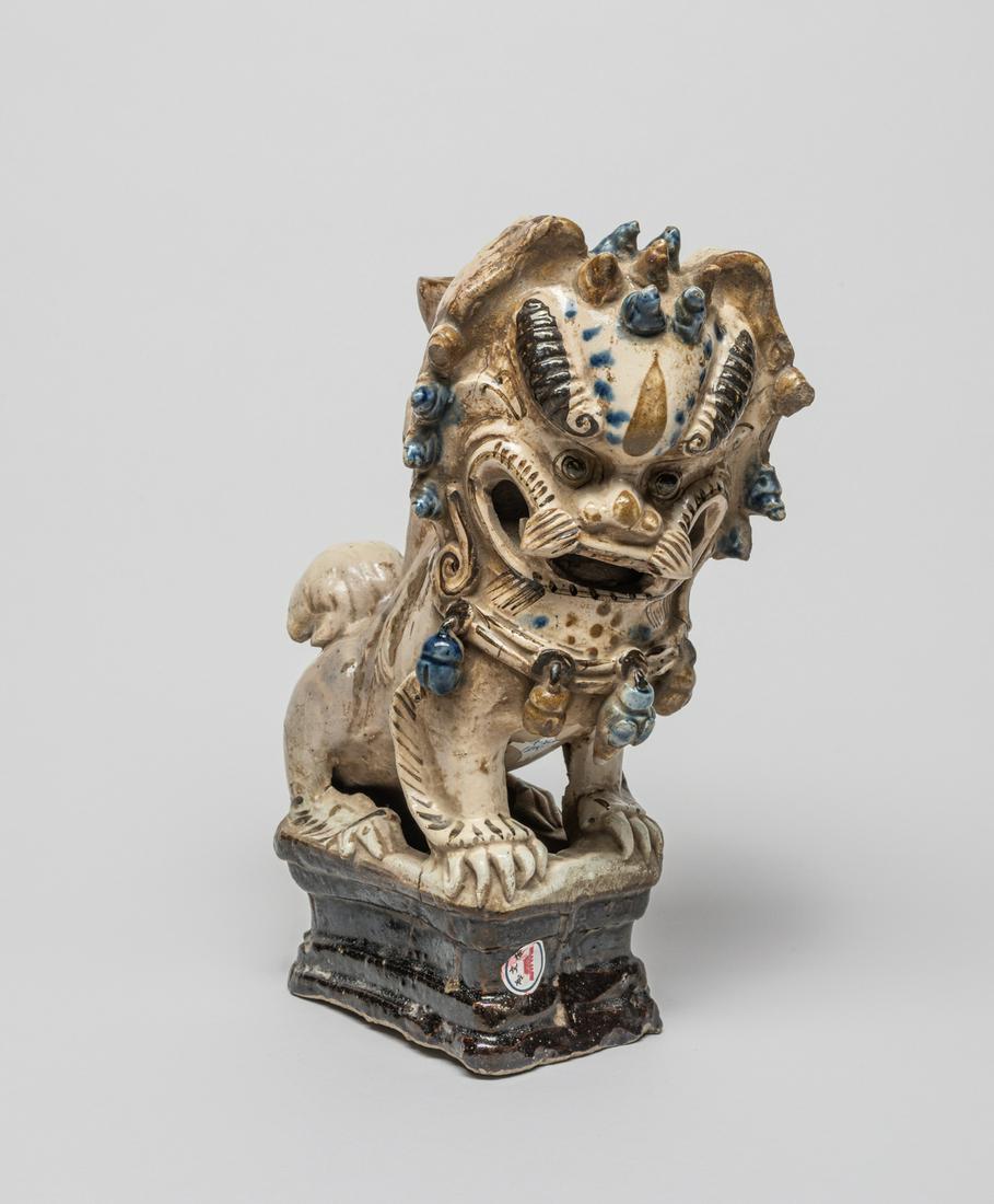 Chinese Stoneware Porcelain Foo Dog: The foo dog crouching on a rectangular stand with its head turned slightly to the right, detailed with bulging eyes, raised spiral horns and curled hair, wearing a string of bells on the neck. 10.5 in