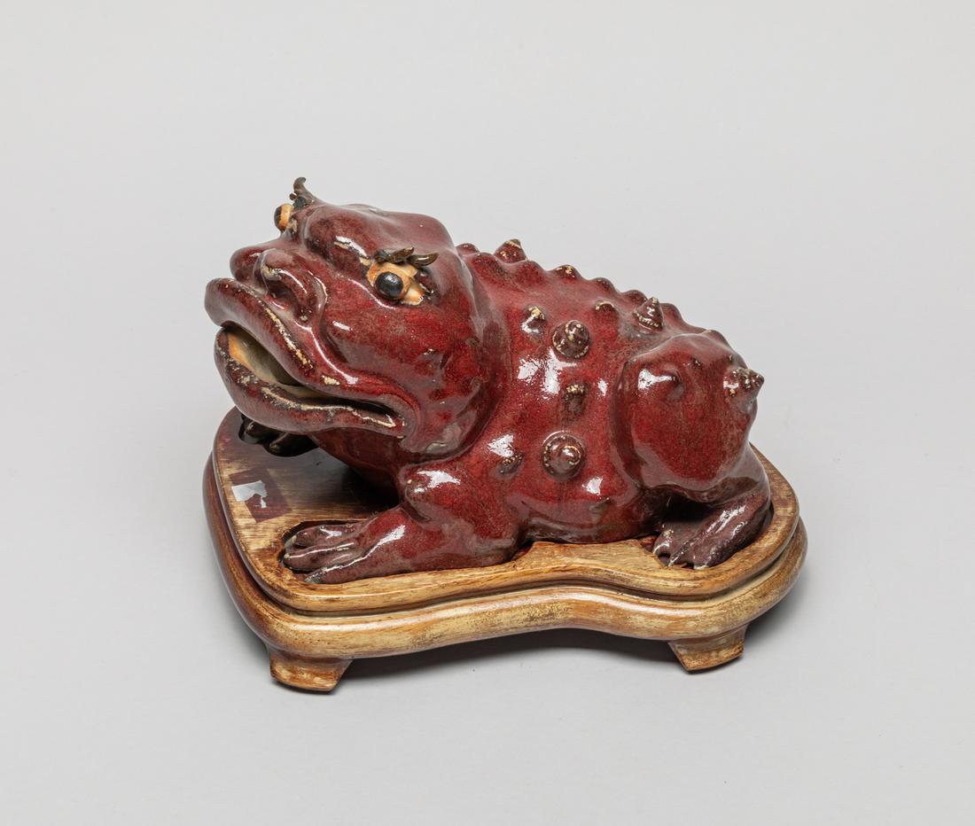 Chinese Flamble Glazed Porcelain Sculpture: Modeled in a recumbent beast form, the body with raised spirals, detailed with bulging eyes and open mouth holding ball, applied with a dark red glaze, With a wood stand. 8.5inch L, 6inch W, 5.1inch H