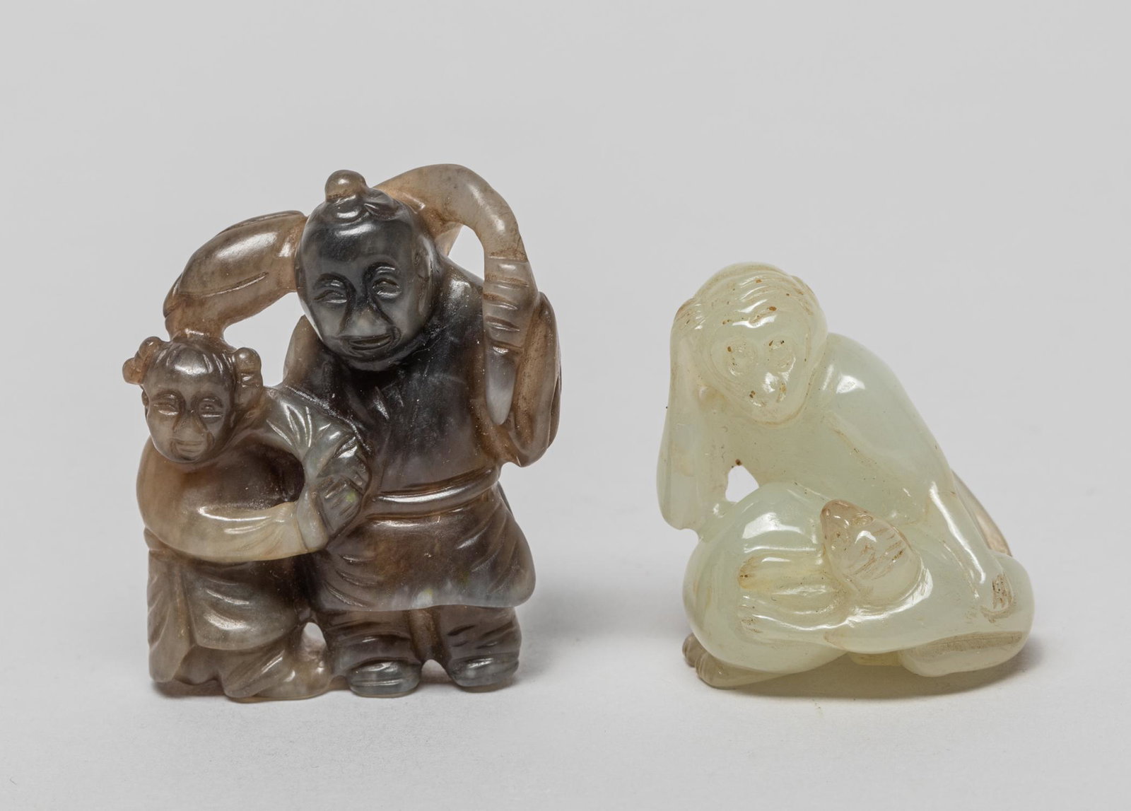 Chinese Manchu Type Jade Toggles: The white jade toggle carved a crouching monkey with its young and a large peach, the black jade toggle carved as a man and a boy standing together and hands grasping a lotus bud with long stem. 1.6in