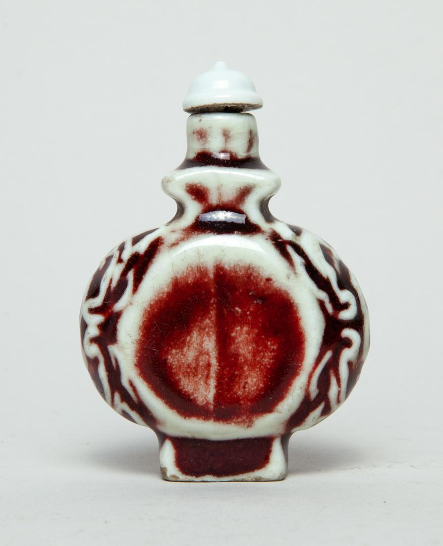 Chinese Red Glazed Porcelain Snuff Bottle: Of ovoid form, the flanks carved with coins and masks, applied with a glaze in dark red and white colors, with a matched stopper. 3.05inch H, 2.15inch L, 1.1inch W.