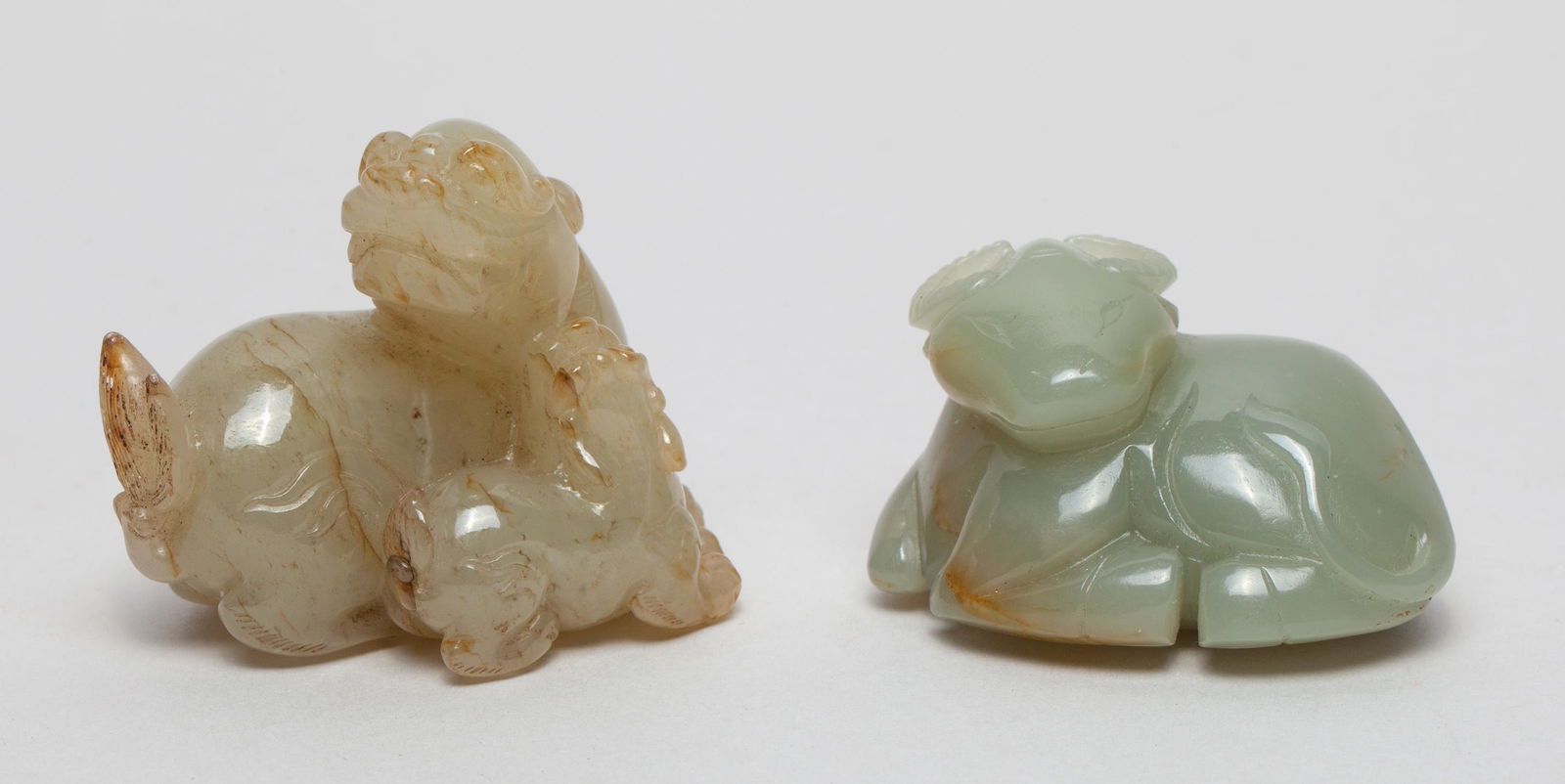 Chinese Jade Toggles of Beast: Including a russet jade beast with its head turned backwards, with its cub nuzzling the parent, and a celadon jade recumbent buffalo with its head turned to the left, the four legs tucked beneath its