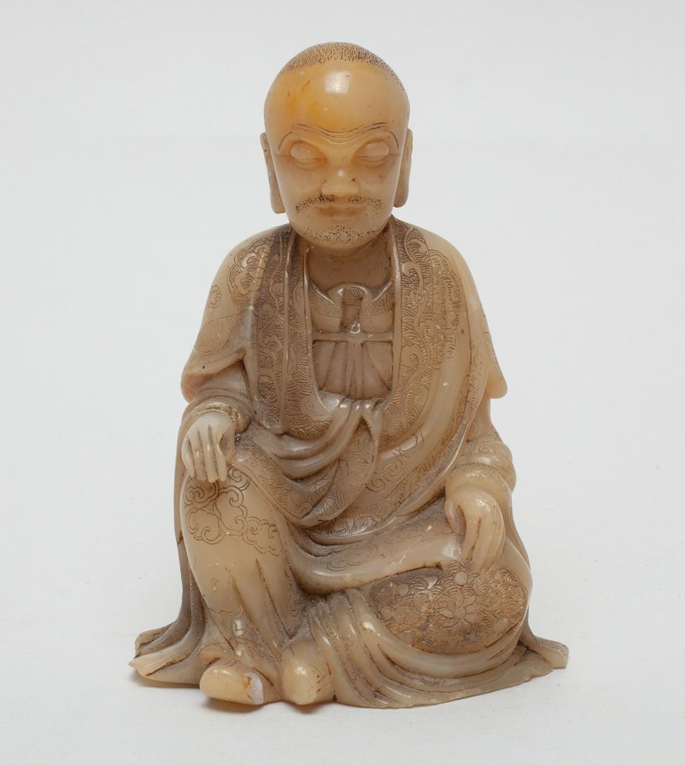Repaired Chinese Shoushan Stone Luohan: Shown seated sculpture, the luohan depicted with a serene expression defined by downcast eyes and a short beard and moustache between pendulous ears, his left hand on his lap, his layered robes engrav