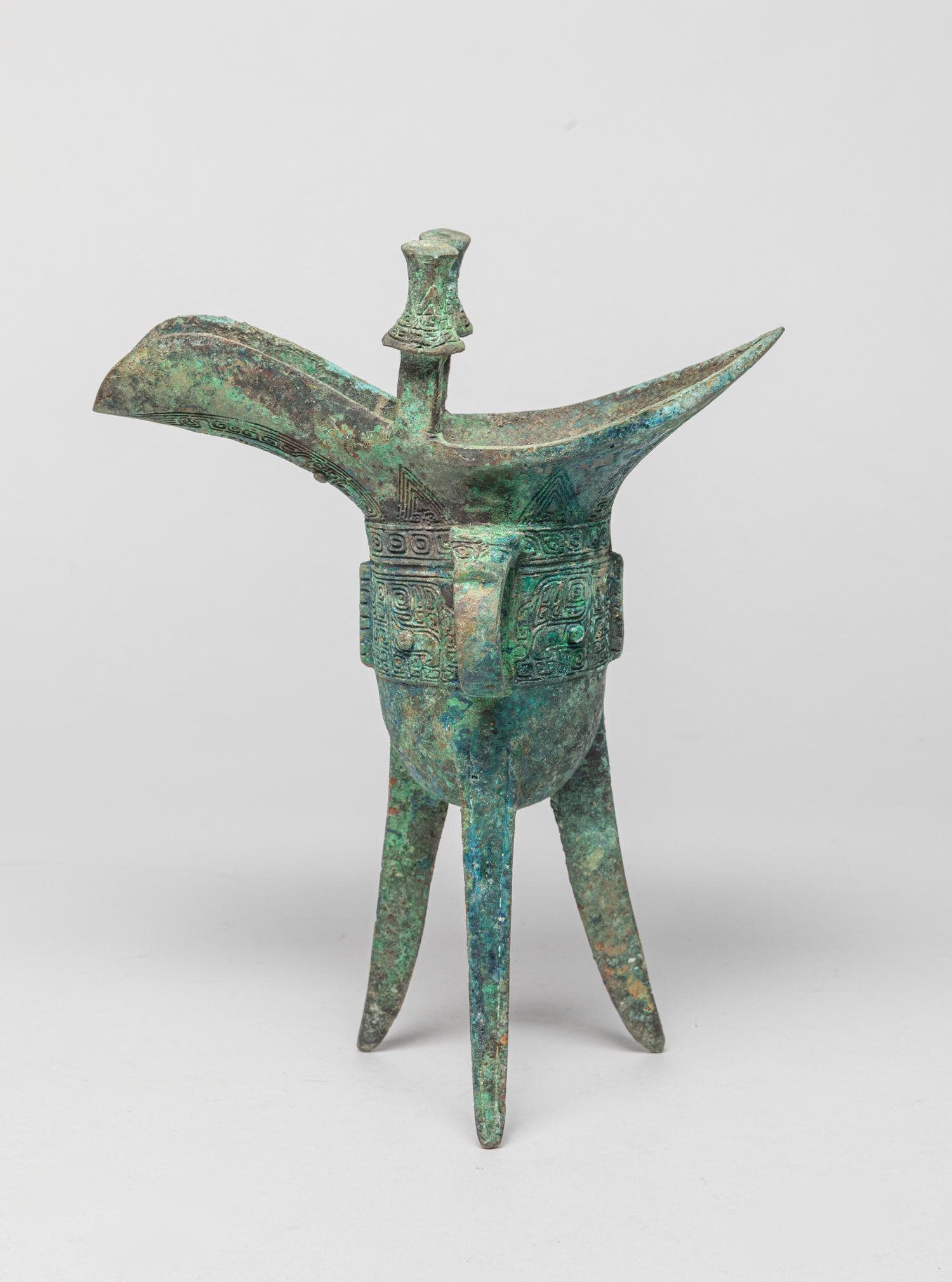 Chinese Bronze Tall Cup: The deep U-shaped body rising from three splayed triangular blade legs to a pointed rim opposite a guttered spout with a pair of posts, the exterior cast with taotie masks divided by three vertical fl