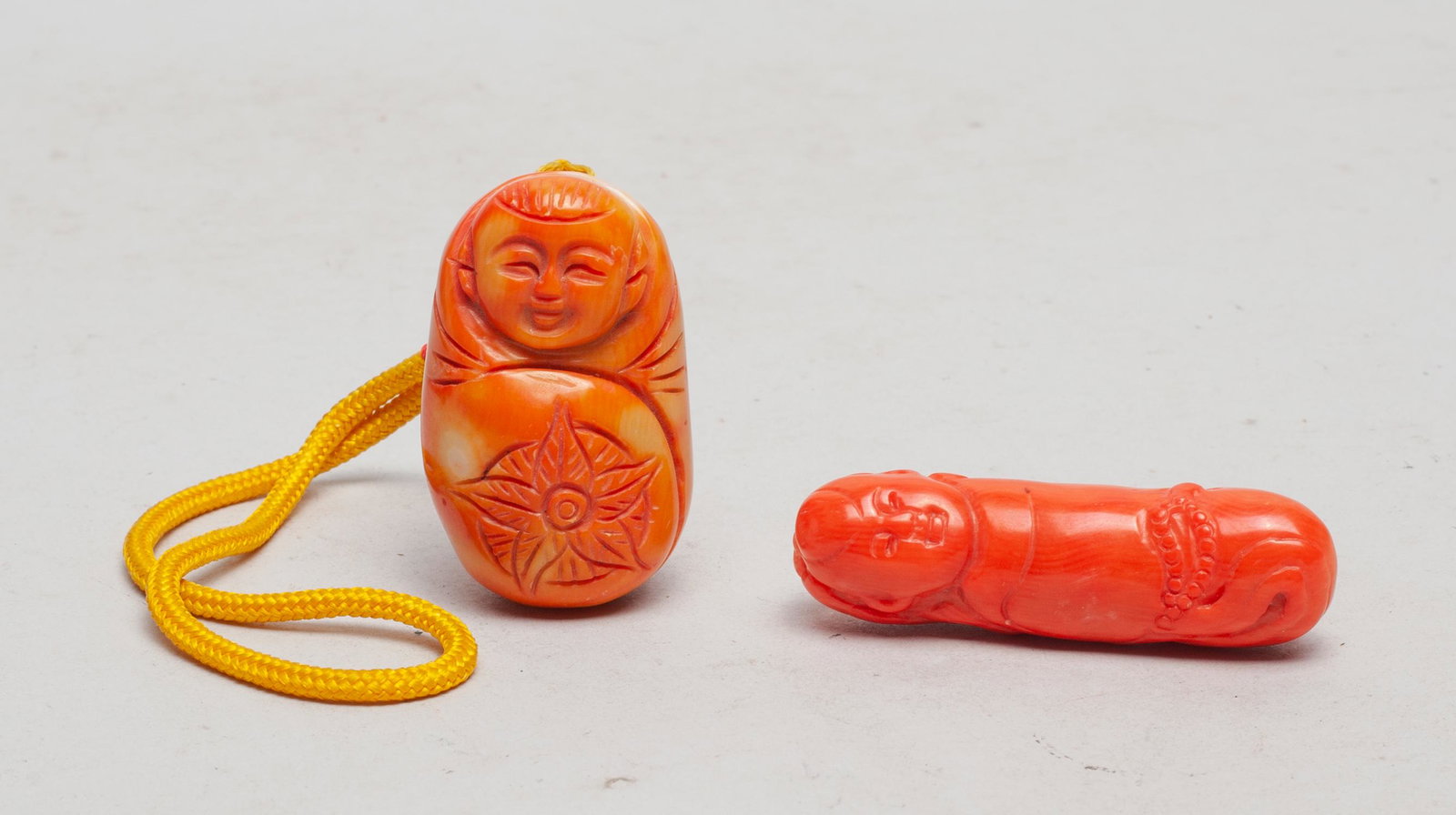 Chinese Coral Like Carvings: One carved as a Buddha and a bat, the other as a laughing boy with a flower, suspended on a white bead. 1.95inch H, 1.2inch L, 1.05inch W (larger).