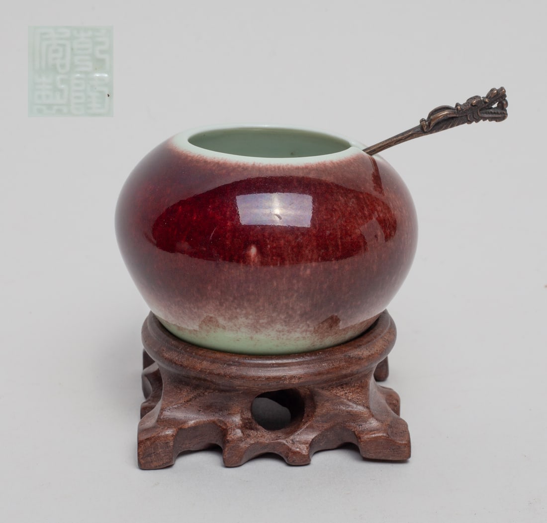 Chinese Porcelain Water Pot: Of ovoid form, the exterior applied in streaked red glaze, the bottom carved with a four-character mark, with a metal spoon and a wood stand. Approx. 2.6-1.7 inch (w/o stand).