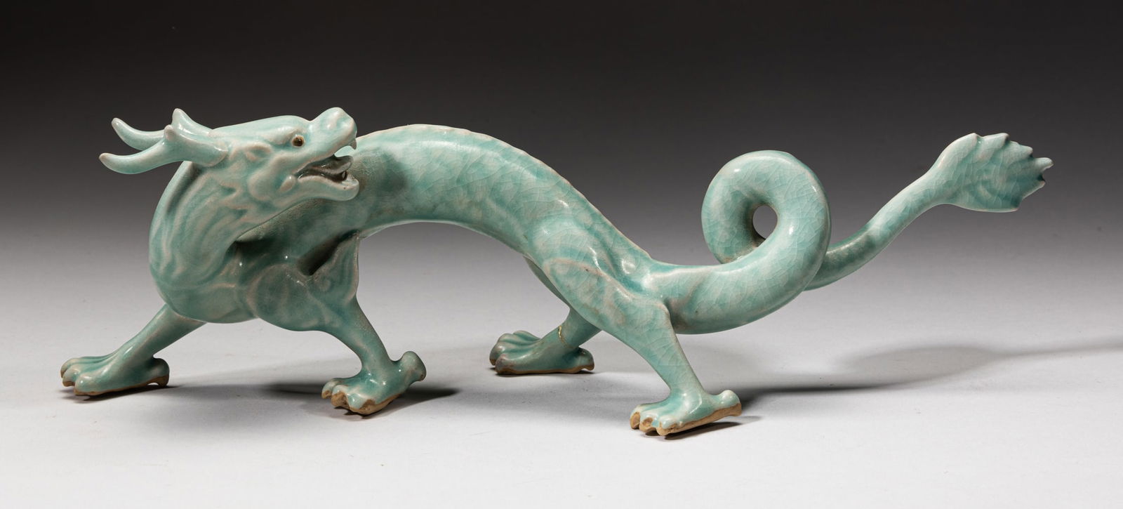 Korean Celadon Porcelain Dragon Stand: One Korean vivid dragon sculpture, glazed in celadon color suffused with crackles, the dragon in walking stance with back raised up and looking back to its spiral tail, with one leg repaired. Approx.