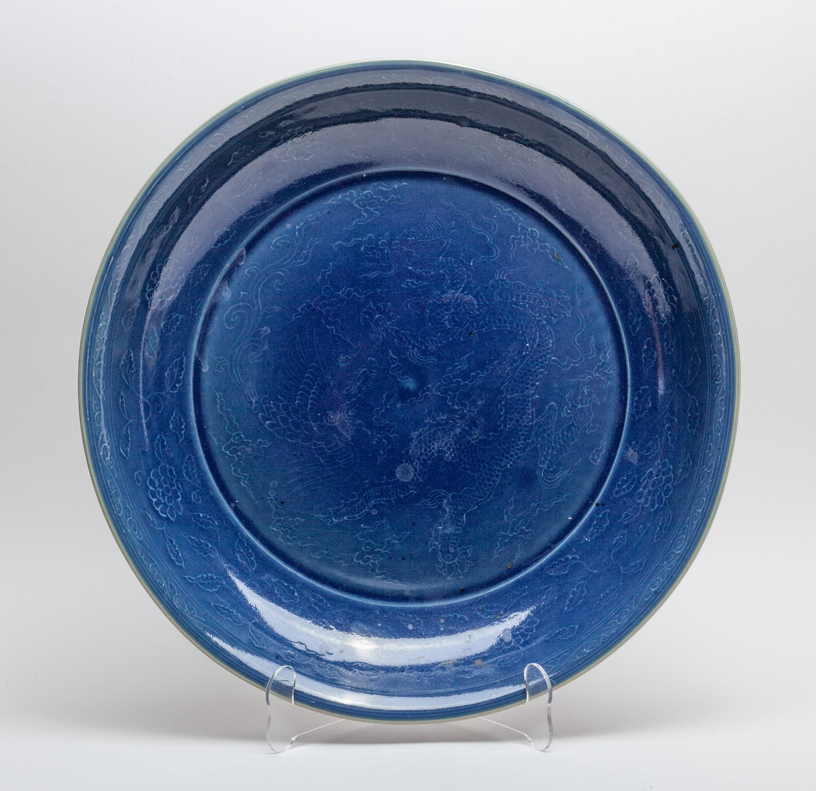 Chinese Blue Glazed Porcelain Charger: The charger interior center carved in low relief with a dragon and a phoenix amidst clouds, the sides with meandering leafy lotus sprays, covered in rick dark-blue glaze save for the unglazed bottom. 