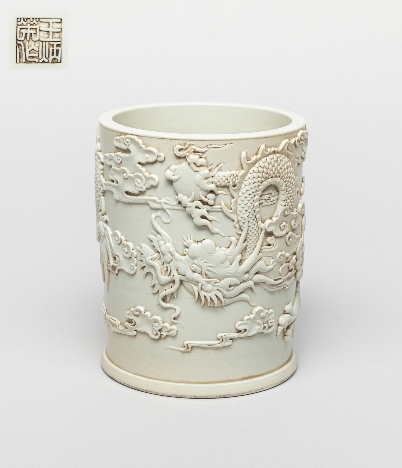Chinese Blanc De Porcelain Brush Pot: Of cylindrical form, the exterior carved in raised relief with a ferocious dragon in pursuit of a flaming pearl amidst clouds, overall glazed in creamy white color, the bottom incised with a seal mark