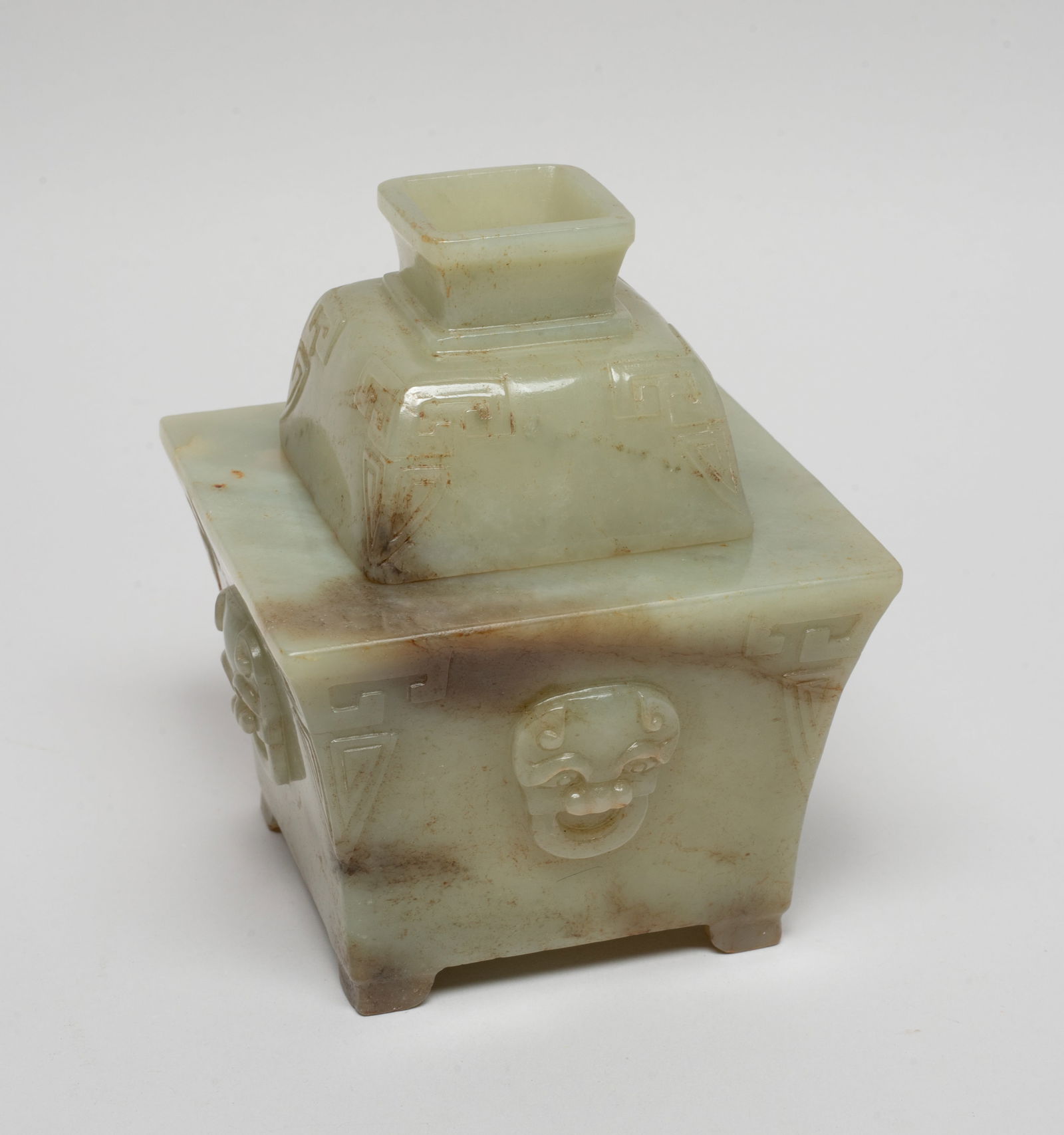 Chinese Jade Covered Cabinet Censer: Of square section, each side carved in raised relief with a beast mask, the body and lid edges carved with stylized leaf motif, the stone of pale celadon tone with russet inclusions. Approx. 4.5*3.6*3