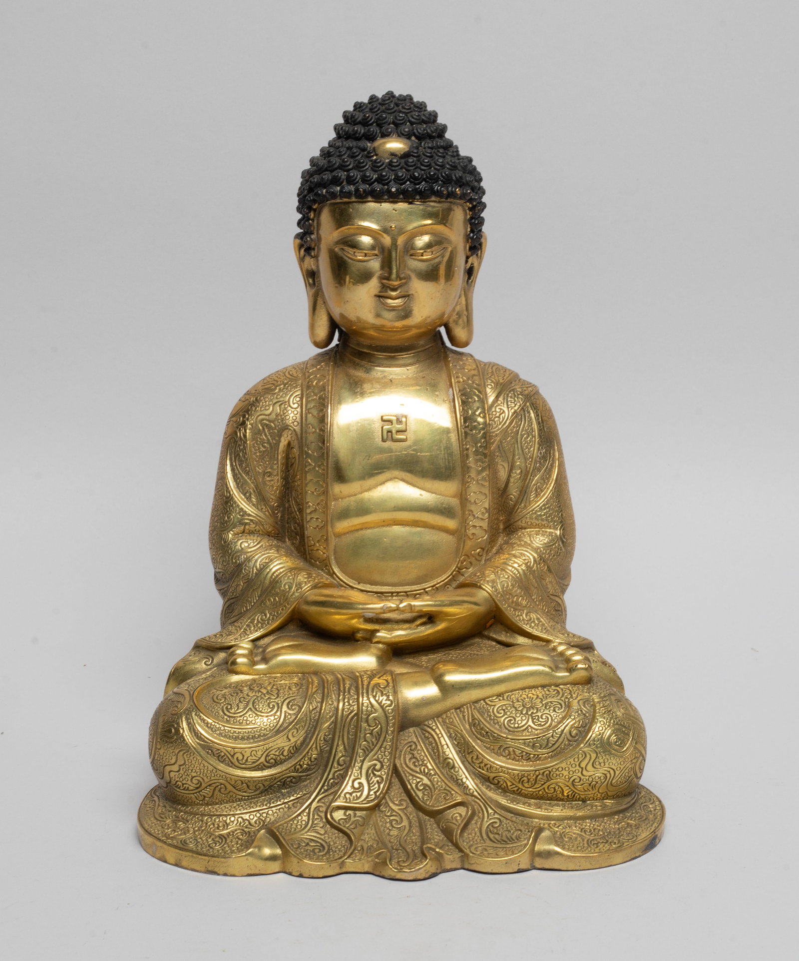 Large Chinese Gilt Bronze Sculpture of Buddha: The seated Buddha wearing a loose-fitting robe open at the chest revealing a swastika symbol, the robe carved with lotus and traditional symbols on circle ground, the hands in dhyana mudra, the serene