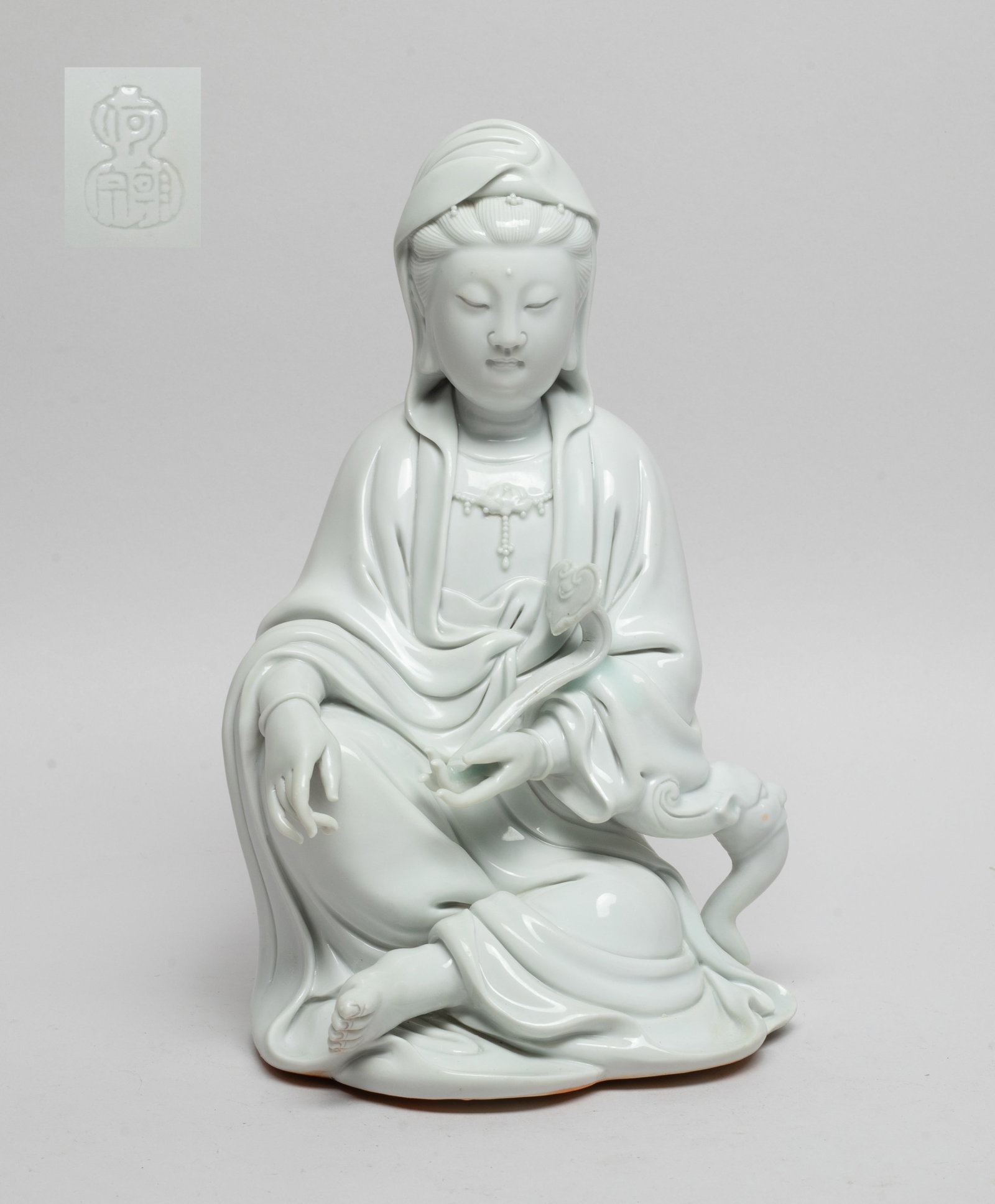 Chinese Blanc De Porcelain Sculpture of Kuanyin: The seated Kuanyin wearing a long flowing robe open at the chest revealing a lotus necklace, her right hand holding ruyi, the exterior covered in even white glaze, the back side marked with four chara
