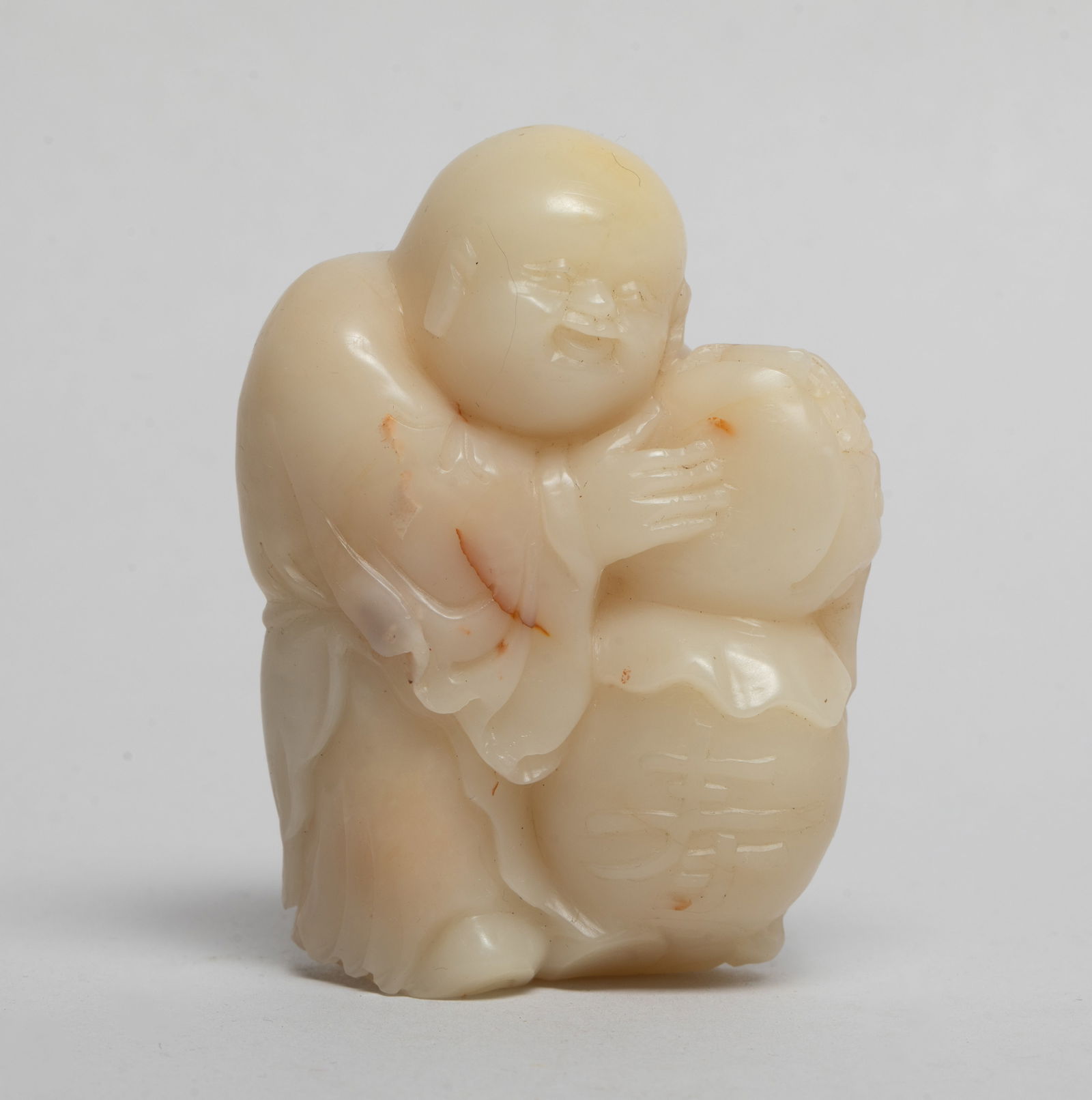 Chinese Shoushan Stone Sculpture of Figure: Carved as a figure holding large peaches inscribed with a Shou character, detailed with a bat and a coin on his back, the stone of beige tone with russet inclusions. Approx. 2.9*2.15*1.07 inch.