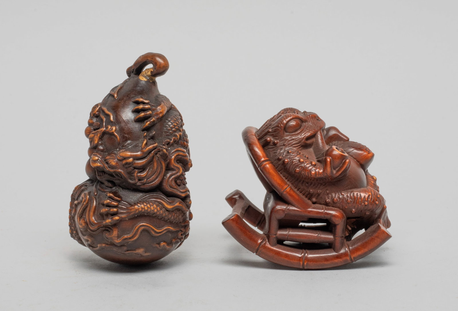 Two Japanese Wood Carved Toggles: One carved as a frog sitting on rocking chair, one webbed foot holding a pipe and one foot holding a duck, the other carved as a dragon encircling a double gourd, borne on short stem issuing smaller g