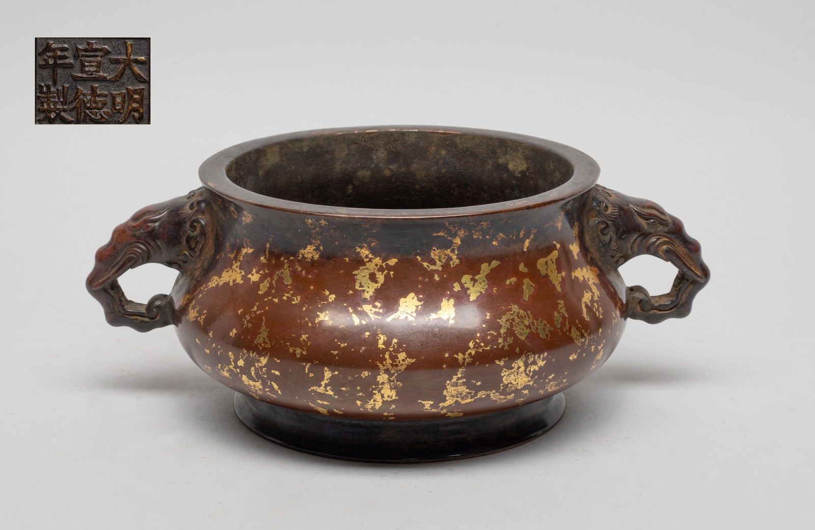 Estate Chinese Gilt Bronze Censer: The compressed body rising from a splayed foot to a broad waisted neck, flanked by a pair of elephant head handles, the mottled brown patina spotted with irregular patches of gilt, the bottom carved w