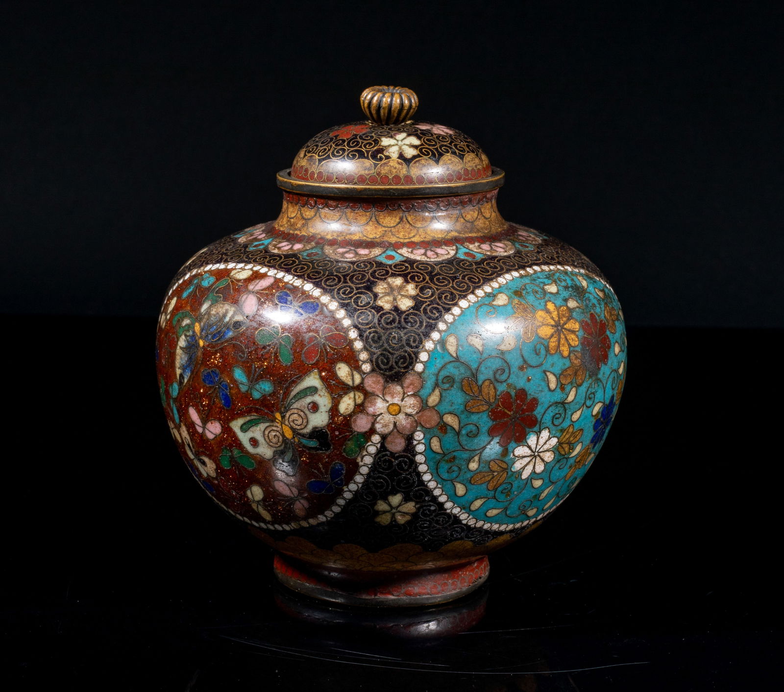Japanese Meiji Cloisonne Covered Censer: The ovoid body decorated with four panels enclosing flower scrolls, butterflies, or a phoenix amidst clouds, against black scroll ground interspersed with flowers, between bands of overlapping petals.