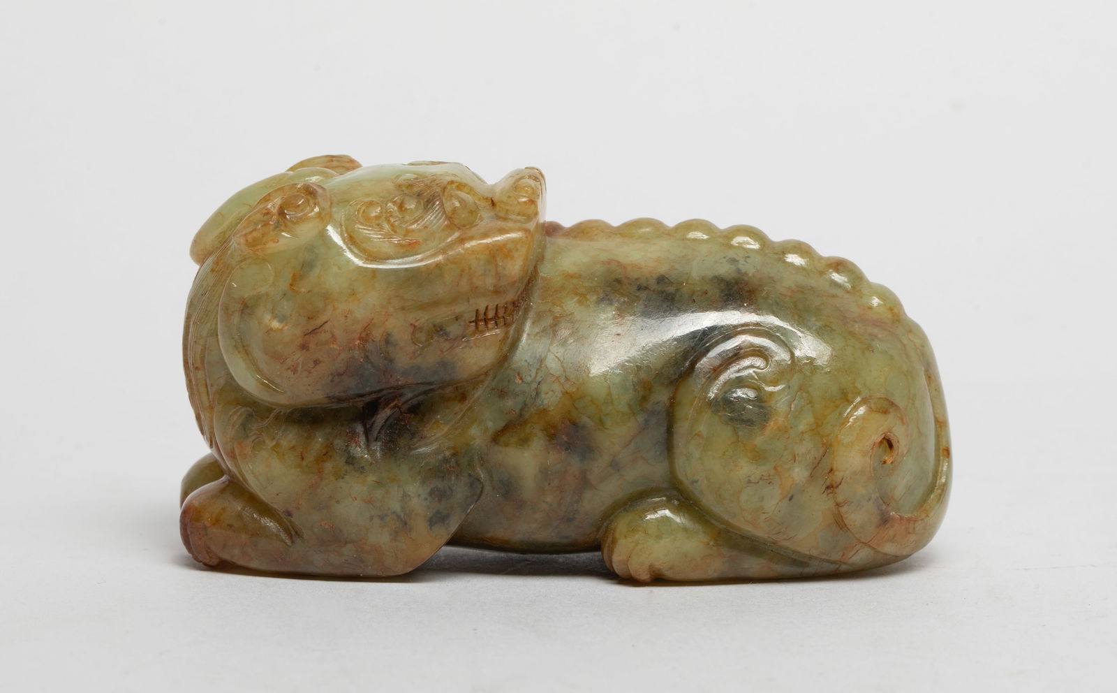 Chinese Russet Jade Sculpture of Beast: The mythical beast powerfully and compactly carved with its head coiled over the left shoulder, the legs tucked underneath the body, the flattened head crowned with a single horn, the jaws open to rev