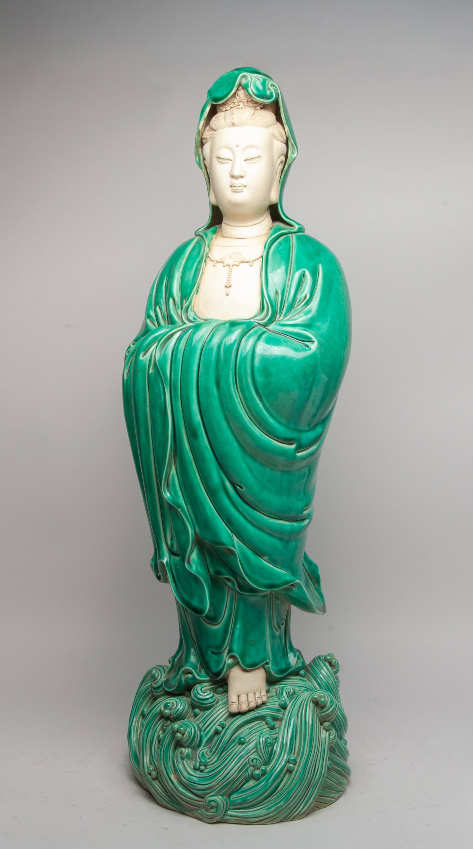 Detailed Chinese Glazed Porcelain Tall Kuanyin: The Kuanyin moulded standing on foaming waves, wearing a long flowing robe covered with crackled green glaze, decorated with a bead lotus necklace, the face with serene expression, the face, chest and