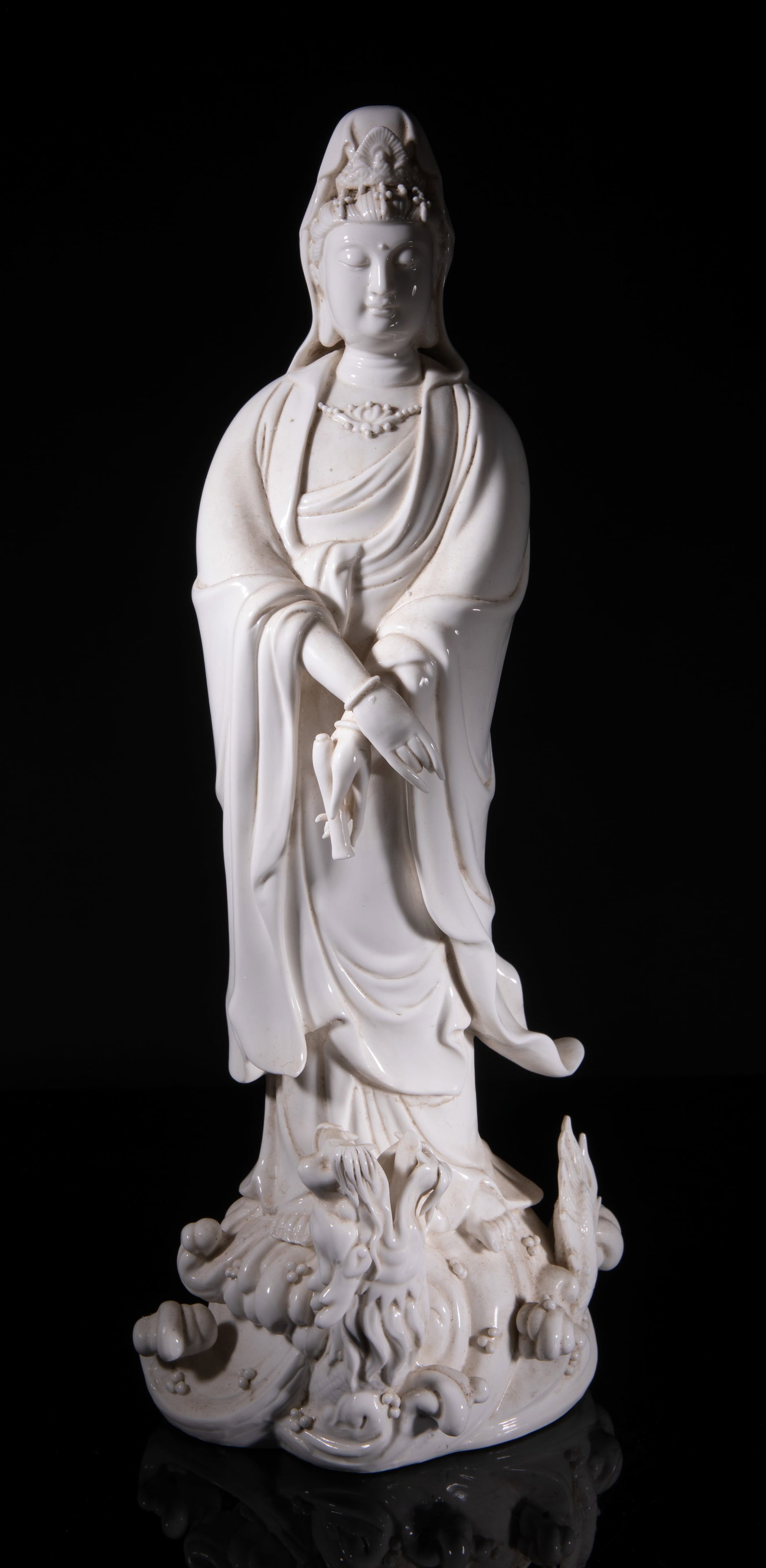 Chinese Blanc De Porcelain Sculpture of Kuanyin: Molded standing on a ferocious dragon among waves, the left hand grasping a bottle with mouth facing down, wearing a long flowing robe open at the chest revealing a lotus necklace, surmounted with a e