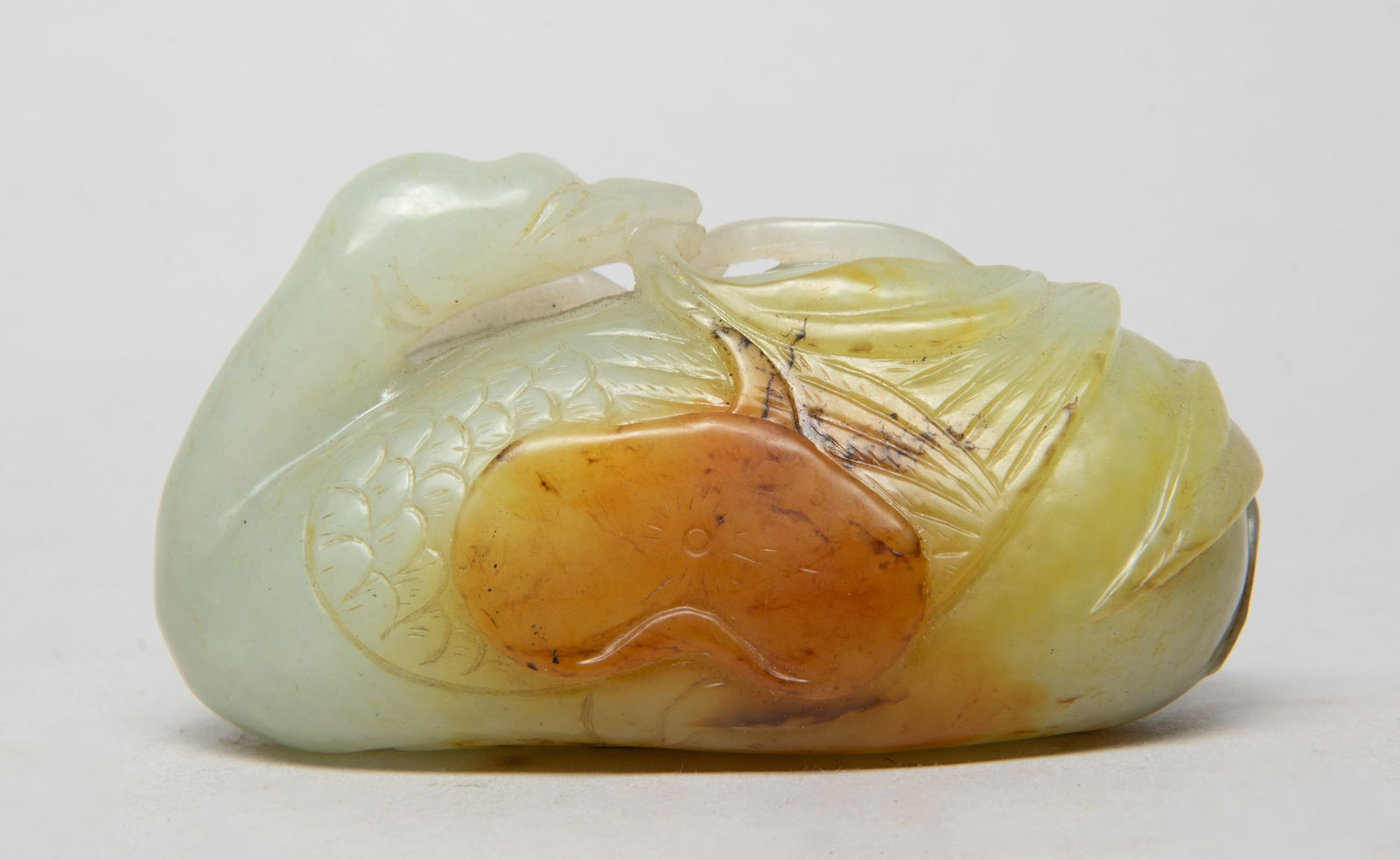Chinese Russet Jade Sculpture of Goose: Carved as a goose with its wings folded with finely-incised feathers and webs tucked under the body, grasping a long stem issuing a lotus leaf in its beak, the stone of pale celadon tone with rich rus