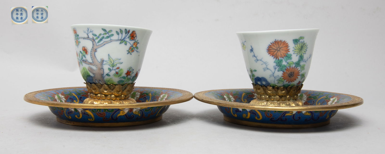 Export Porcelain Tea Cups on Cloisonne Base: One tea cup exterior painted with chrysanthemums and butterflies, the other painted flowers and a pomegranate tree, both inscribed with related poems, marked with six characters on the base, with cloi