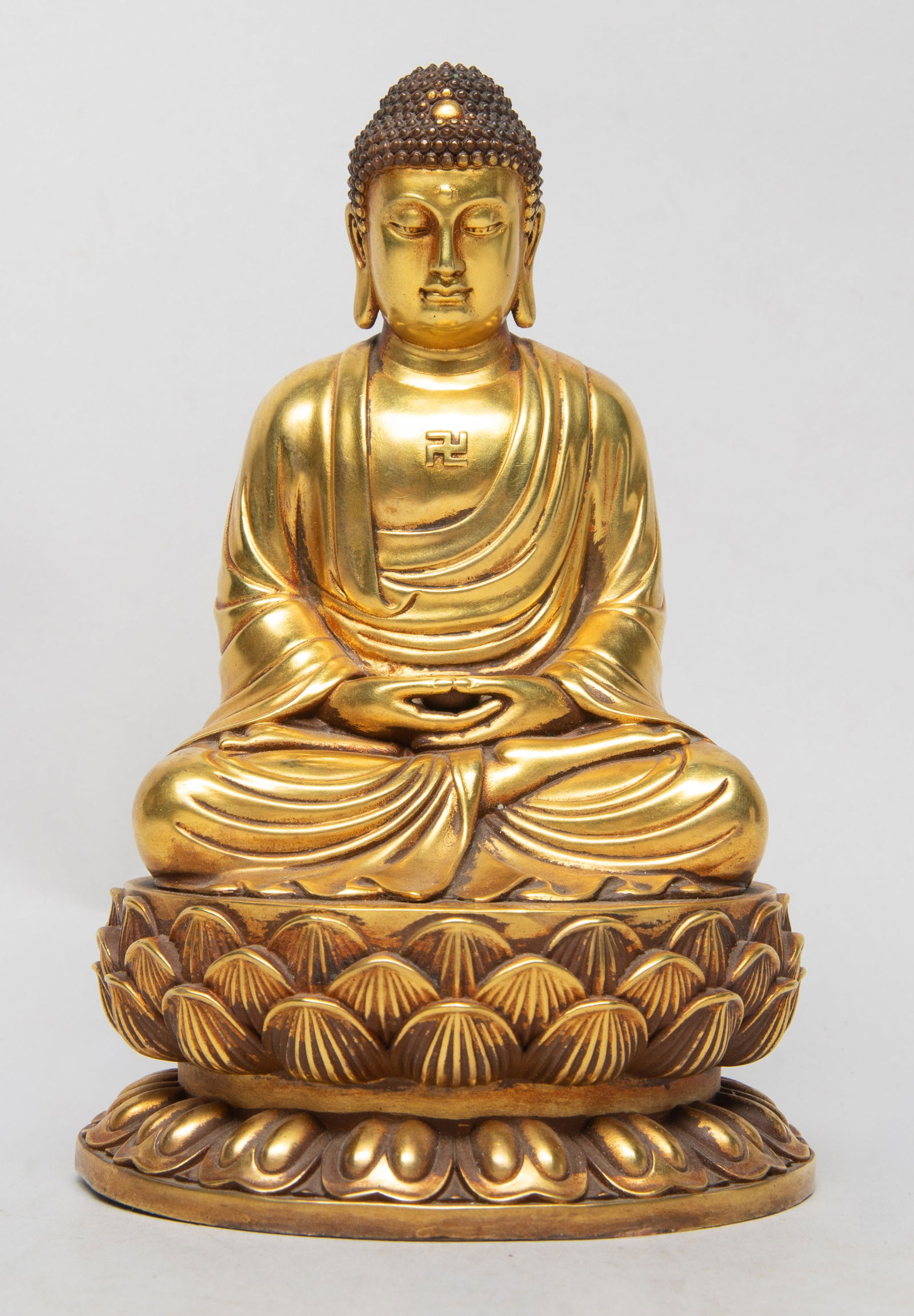 Vintage Chinese Gilt Bronze Sculpture of Buddha: The Buddha cast seated on a waisted lotus plinth, the hands in dhyana mudra holding a bowl, wearing a loose-fitting robe open at the chest revealing a swastika symbol, the face with serene expression