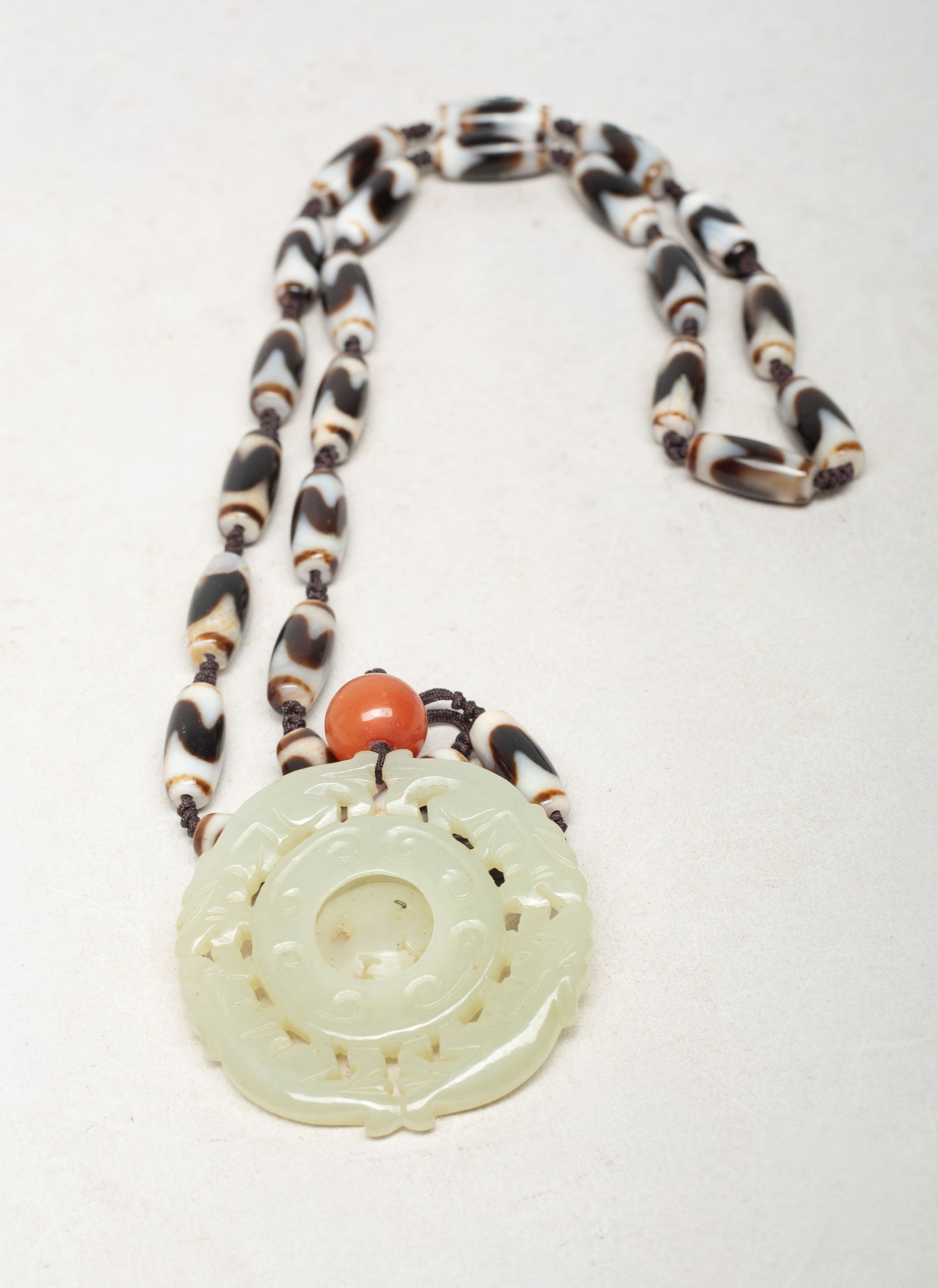 Chinese Dzi Bead Necklace w/ Jade Pendant: Chinese dzi bead necklace, suspended with a red gemstone bead and a jade pendant, carved as two pairs of confronting beasts framing a circular bi with a movable center. Approx. 2.1 inch D (jade pendan