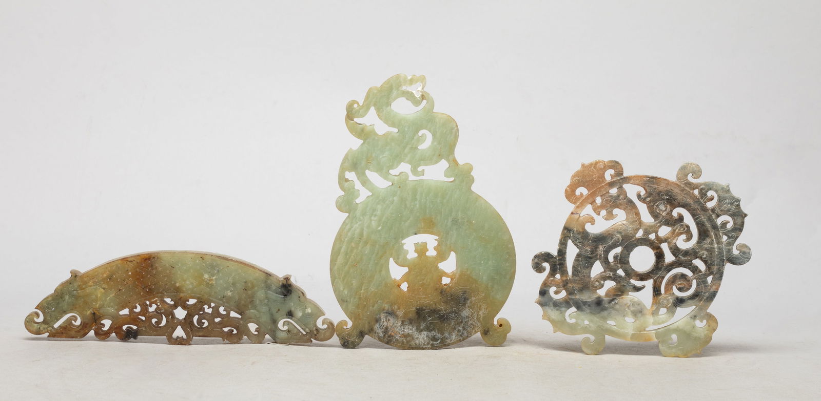 Estate Chinese Export Jade/ Stone Carvings: Three jade / stone carvings, one carved with a dragon head at either terminal, one reticulated with sinuous stylized dragons, the other carved with a beast above a circular disc with a figure at the c