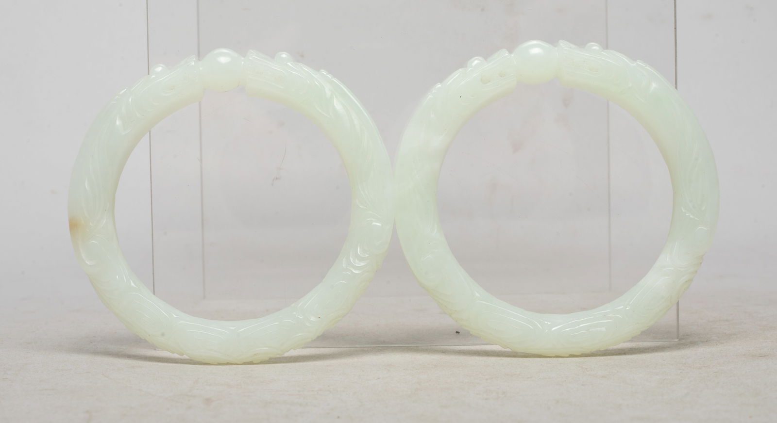 Two Chinese Export Jade Carved Dragon Bangles: Each bangle finely worked in the form of two dragons sharing the same body facing each other at the top, both the mythical beasts fighting over a 'pearl'. Approx. 2.9 inch outer diameter, 2.15 inch in