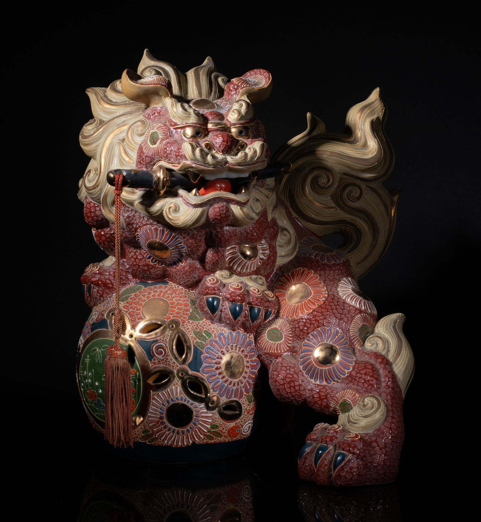 Large Japanese Kutani Ware Porcelain Sculpture: Molded as a mysterious beast carrying a ball in front paws, the mouth agape grasping an attribute suspended with tassel, the body decorated with flowers. Approx. 14.5*12*11.3 inch.