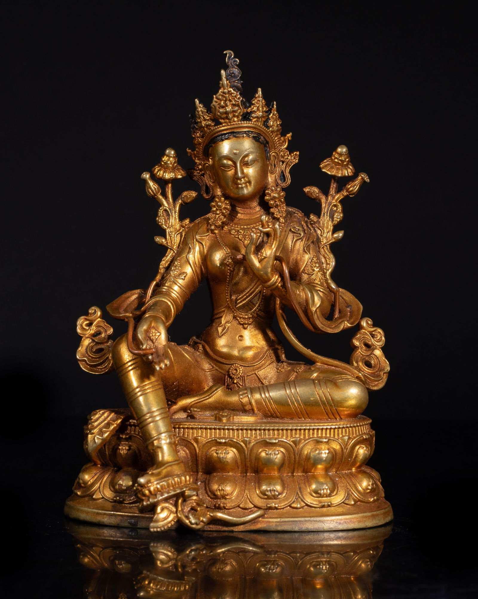 Chinese Gilt Bronze Cabinet Buddha: The Buddha cast seated on a waisted lotus base, the right leg stretching out and stepping on lotus, the hands in elegant mudra holding long stems issuing flowers, wearing elaborate jewelry and diadema