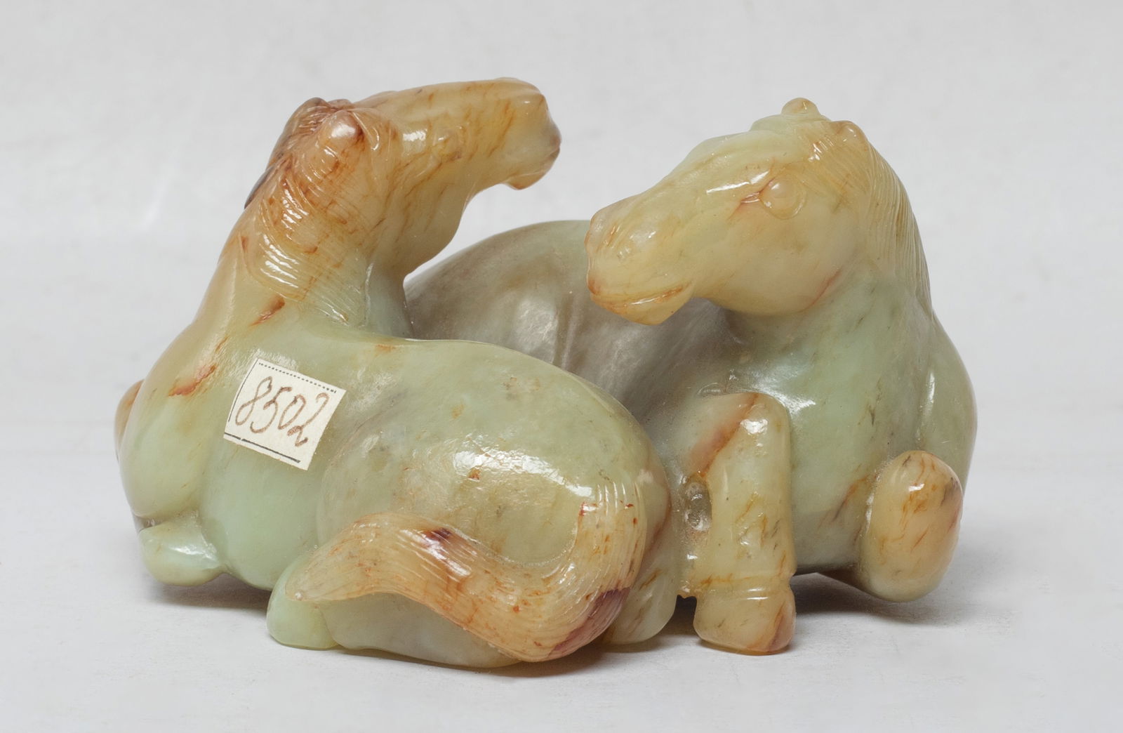 Estate Chinese Russet Jade Sculpture of Horses: The two recumbent steeds posed nestled together back to front and finished on the underside, their heads and manes undercut, carved with russet inclusions in the cloudy gray-green matrix. Approx.