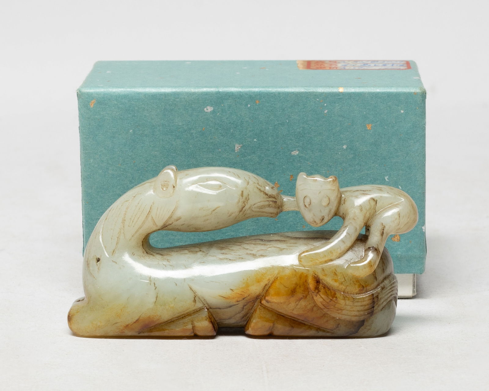 Chinese Jade Carved Sculpture of Horse: The recumbent horse with its head turned to the rear and tail flicked over its haunch, a monkey clambering on its back, the stone of pale celadon tone with russet inclusions, with box. Approx.