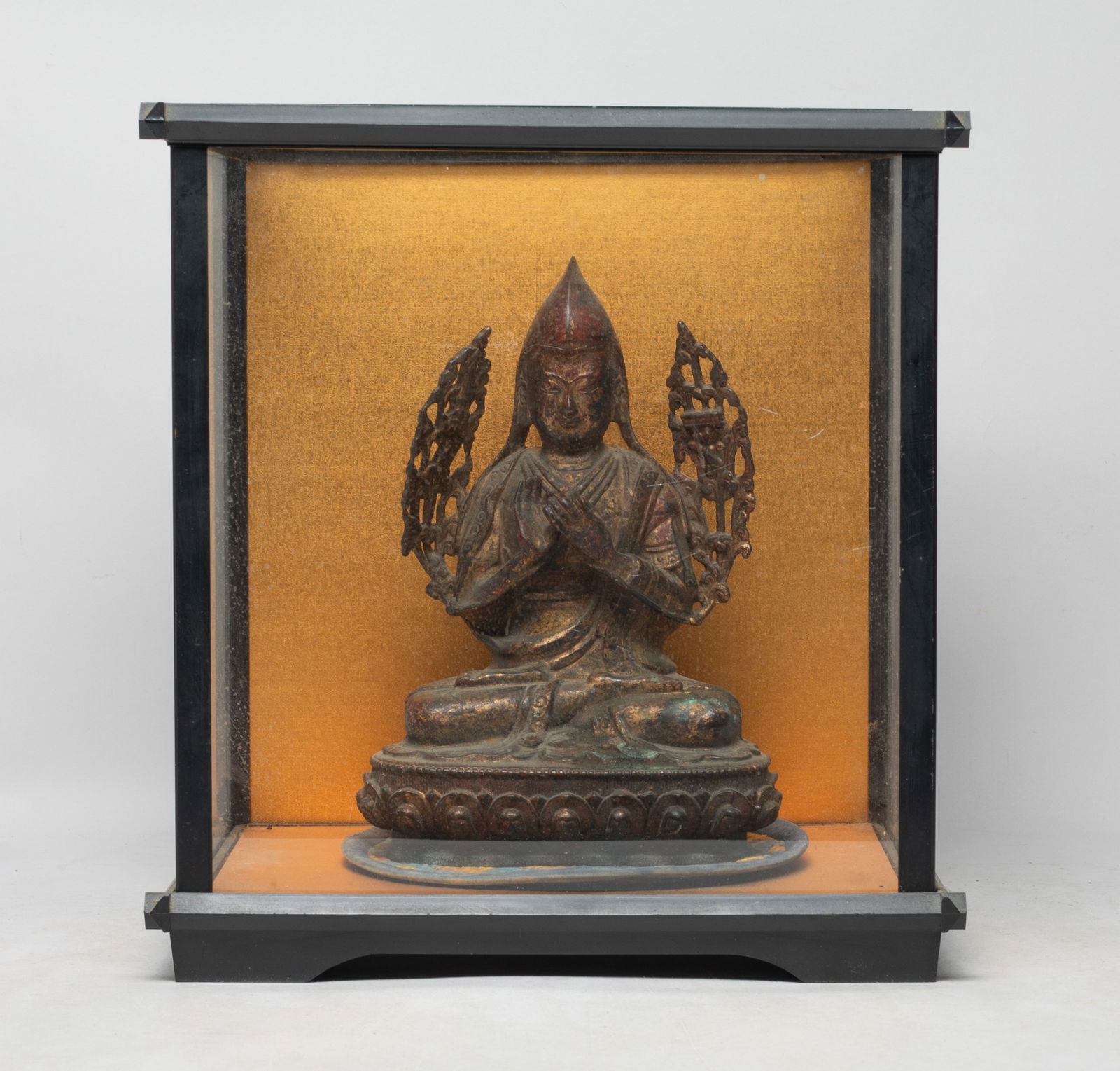 Chinese Bronze Buddha w/ Glass Case: The Buddha cast seated on a lotus base, the hands in dharmachakra mudra, wearing a tall pointed hat, flanked with flowers around the arms, with a glass case. Approx. 11*10.5*8 inch (case size).