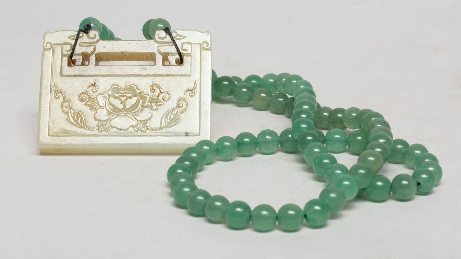 Chinese Export Necklace w/ Jade Lock Pendant: Chinese stone bead necklace suspended with a lock form jade pendant, one side carved with flowers and the reverse inscribed with four characters. Approx. 2.37*1.7 inch (jade size). The Estate of Store