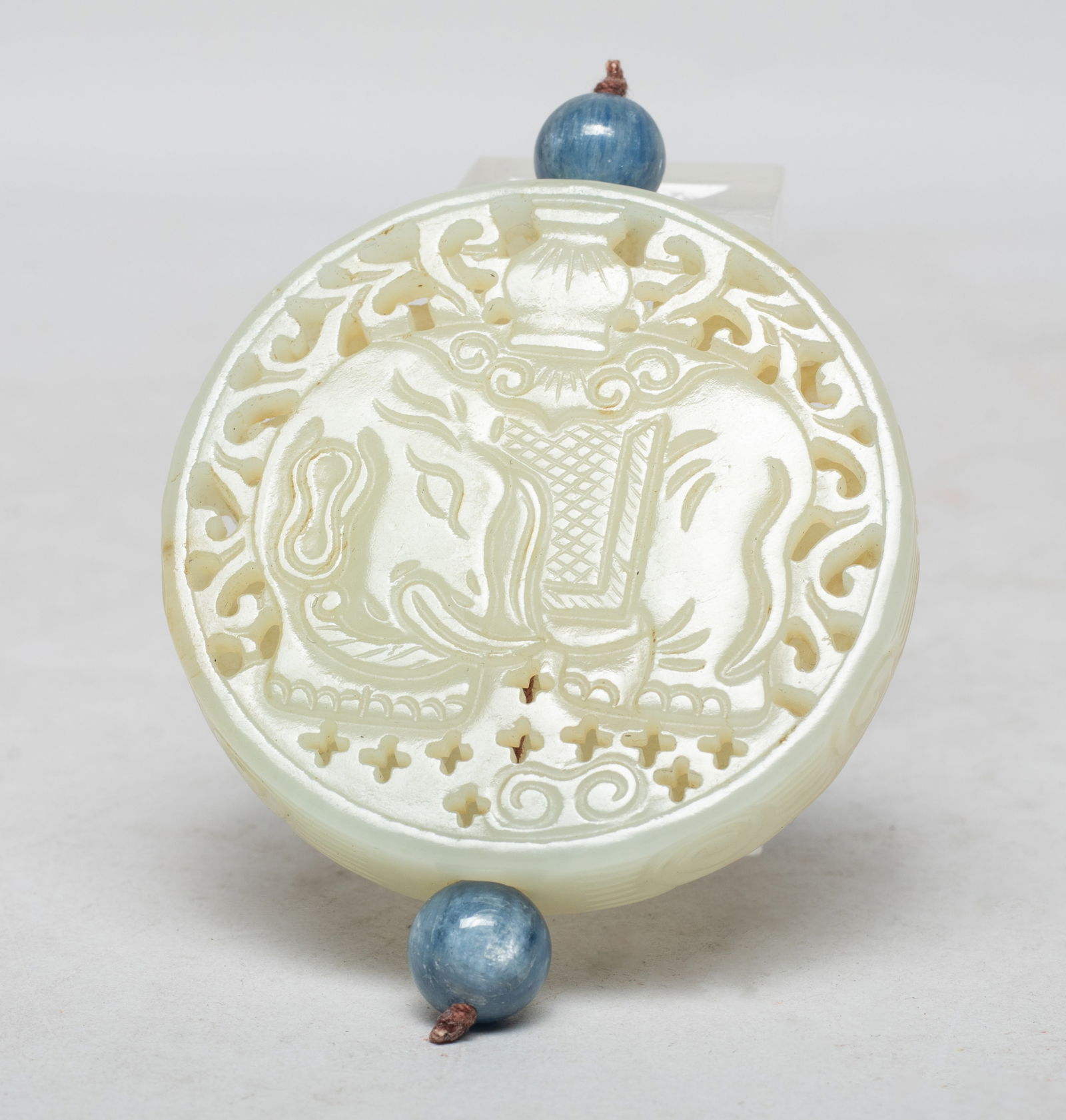 Chinese Export Jade Pendant: Of circular shape, both sides skillfully carved in openwork with a standing elephant holding a vase on the back, the animal head turned backward, with two blue gemstone beads. Approx. 2.15 inch D. The