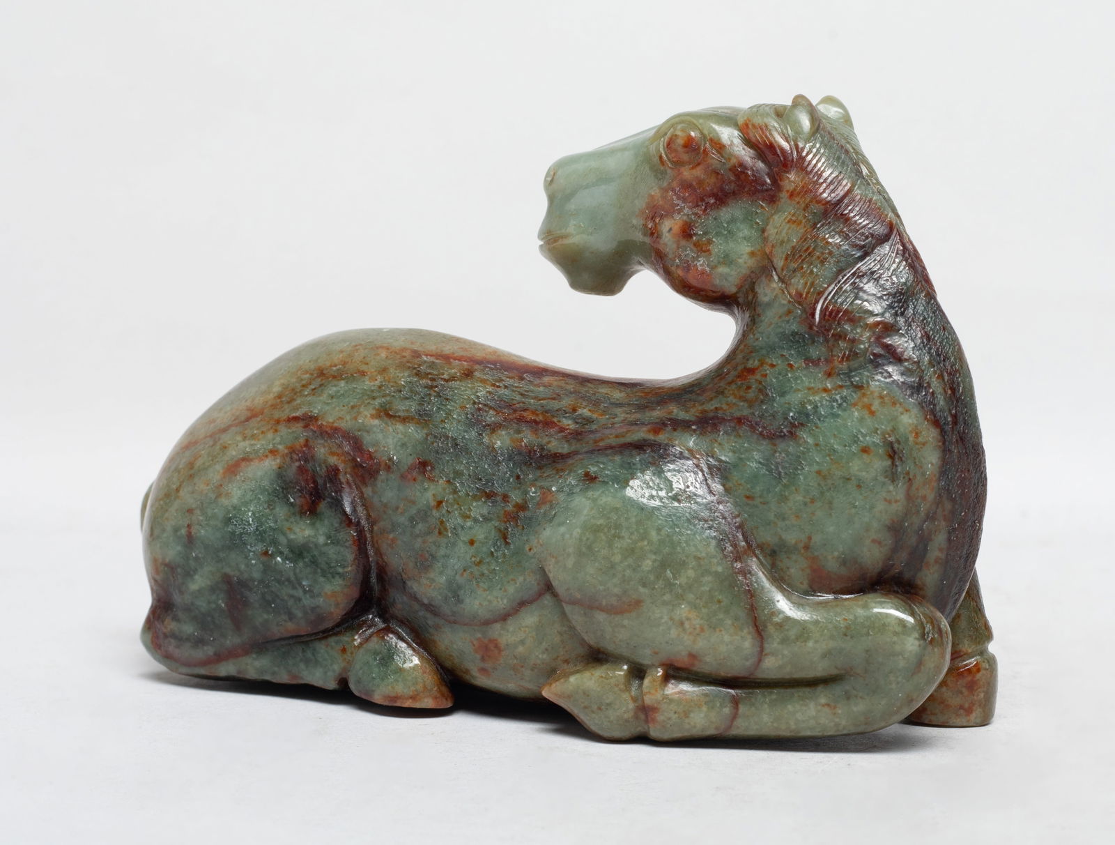 Lg Chinese Russet Jade Carved Sculpture of Horse: The powerful recumbent horse carved with its head turned towards the back, the head with flared nostrils and bulging eyes, below the ears and flowing incised mane, the tail resting along the left flan