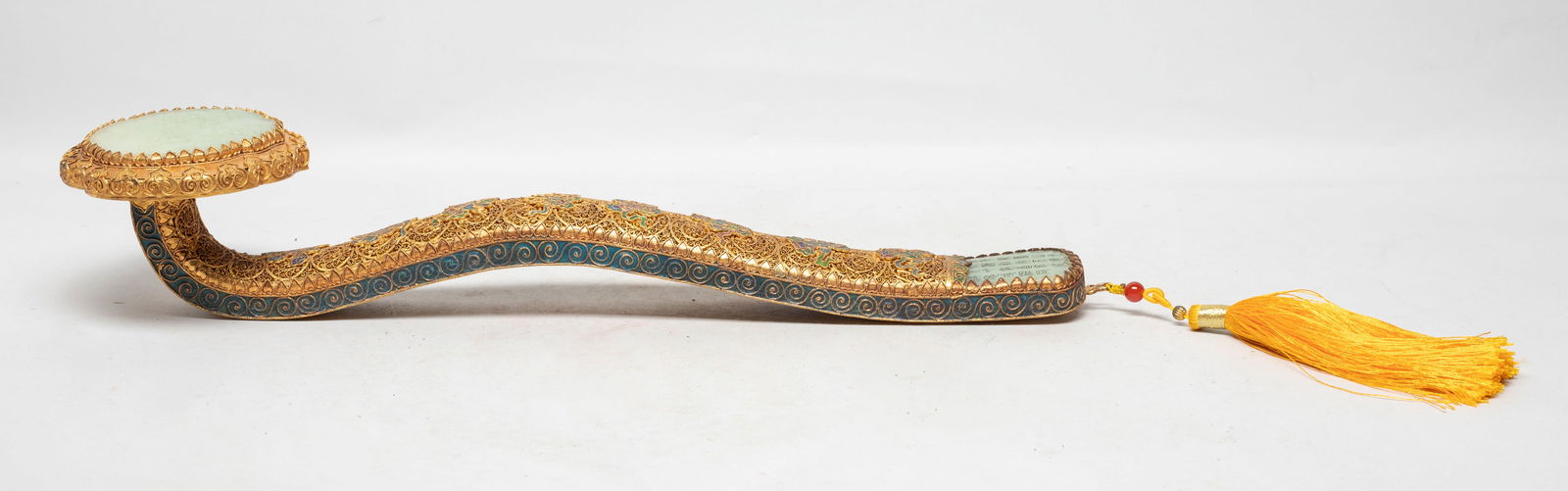 Long Chinese Jade, Gold Color Sculpture of Ruyi: The ruyi head inlaid with a pale celadon jade plaque carved with landscape scene, the end inlaid with jade incised with inscription, the arched shaft enameled on the upper side with Buddhist eight tre