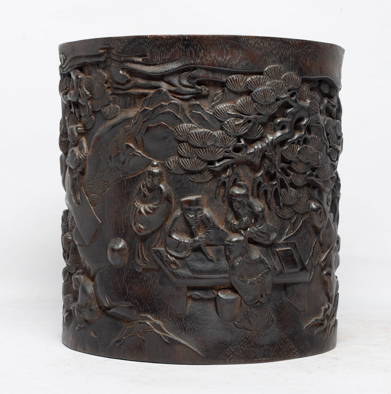 Large Chinese Agarwood Carved Brush Pot: Of cylindrical form, carved with scholars playing instrument, playing chess and viewing paintings in a plantain and pine grove as attendants prepare tea. Approx. 8.1*7.7 inch. The Estate of the Store