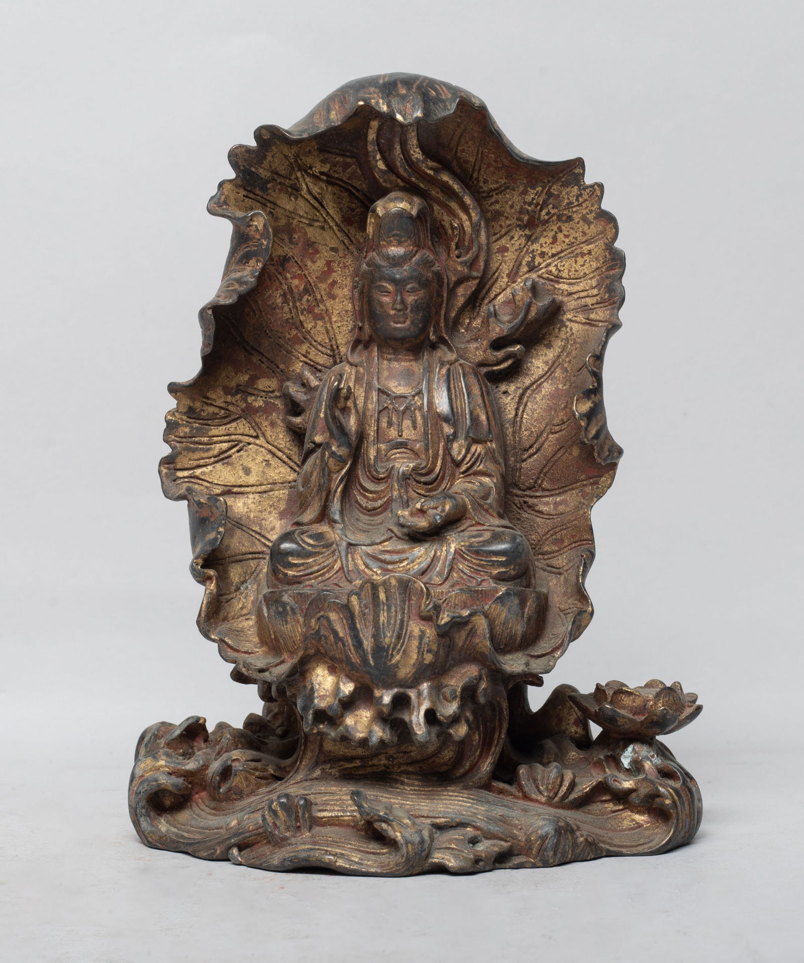 Chinese Gilt Bronze Sculpture of Kuanyin: The Kuanyin cast seated on a lotus throne in front of a large lotus leaf, with a pond of full lotus and rockery below, the left hand holding an implement and right hand in elegant mudra, the back insc