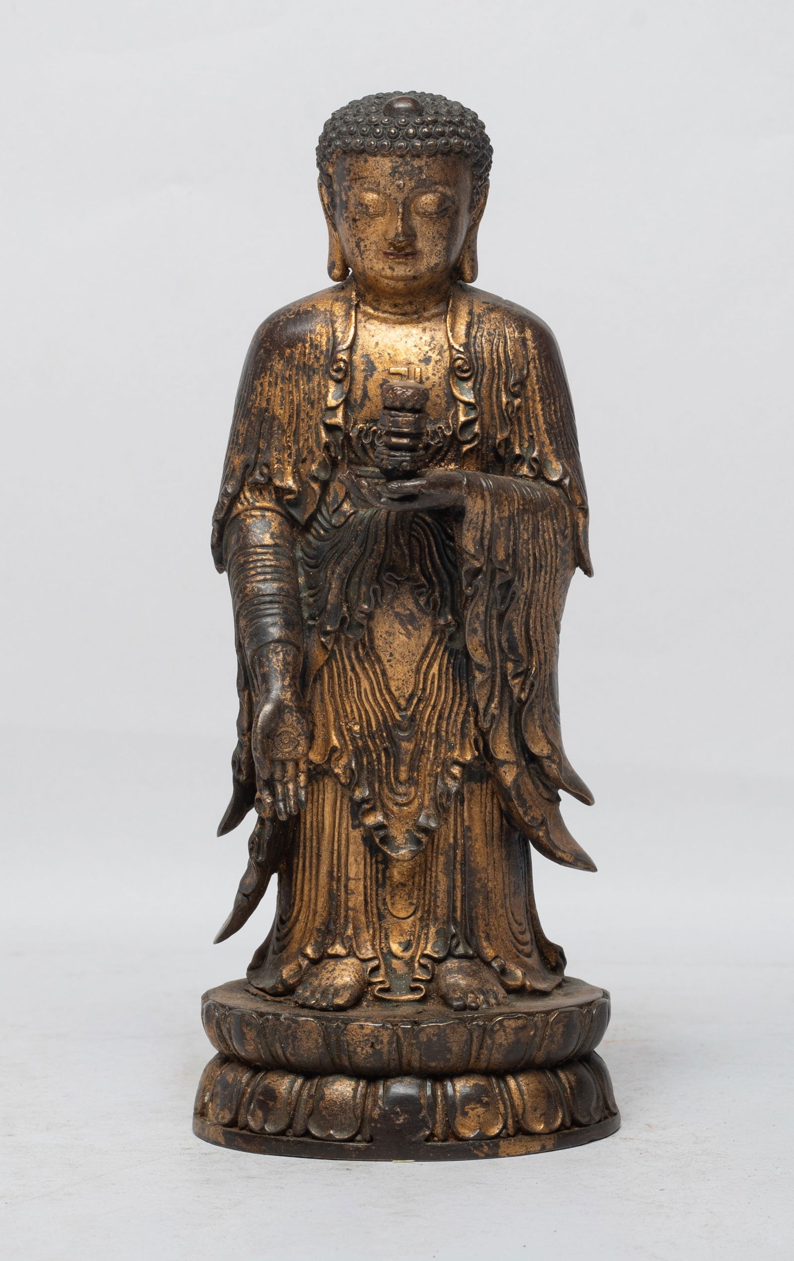 Chinese Gilt Bronze Sculpture of Buddha: The Buddha cast standing on a waisted lotus base, the left hand holding an implement and right hand pointing downward, dressed in long flowing robe open at the chest revealing a swastika symbol. Appro